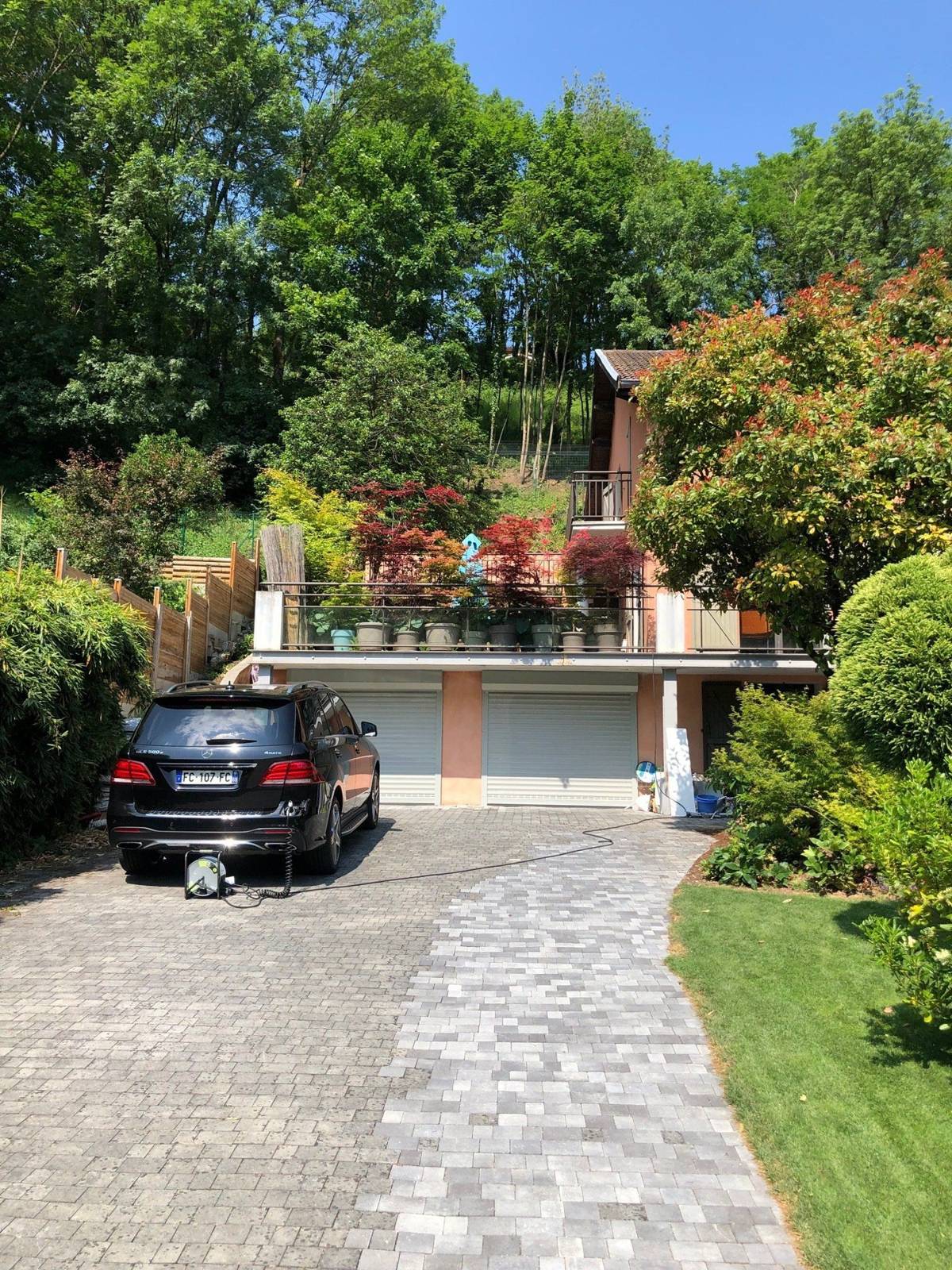 Une allée pavée en pierre grise mène à une maison à flanc de colline avec deux garages, un 4x4 noir garé à gauche et un jardin verdoyant.