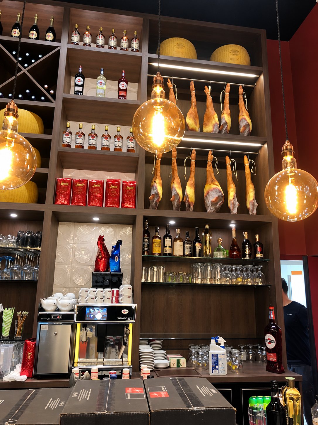 Derrière un comptoir de bar, des étagères en bois sombre présentent des jambons suspendus, des meules de fromage, des bouteilles d'alcool, des verres et une machine à café, sous de grandes ampoules décoratives.