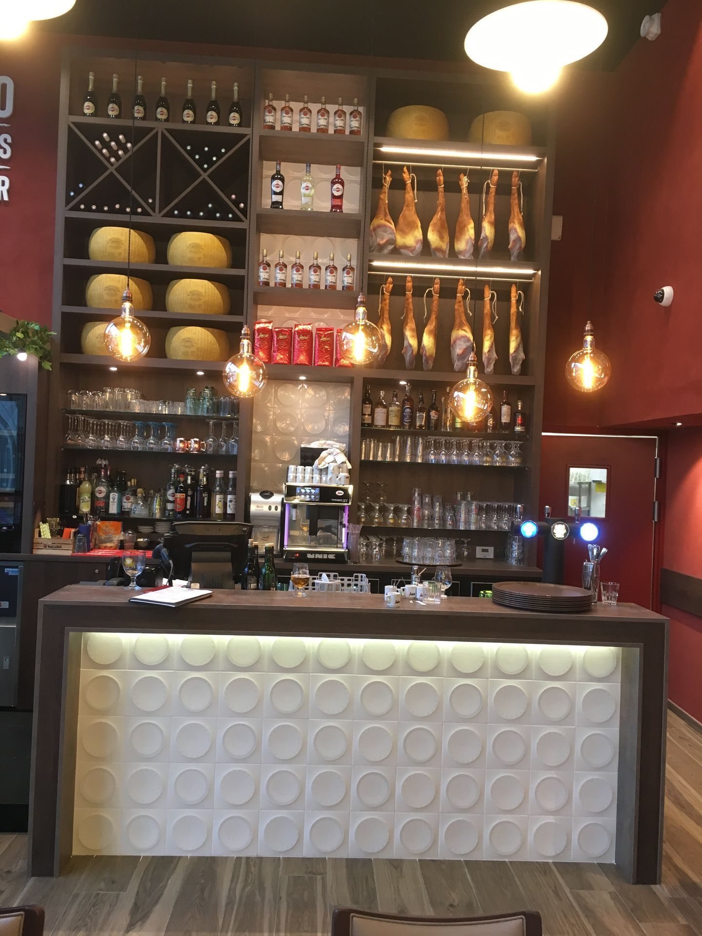 Comptoir de bar blanc éclairé devant une étagère en bois garnie de bouteilles, de meules de fromage, de jambons suspendus et de verres, sous des ampoules à filament.