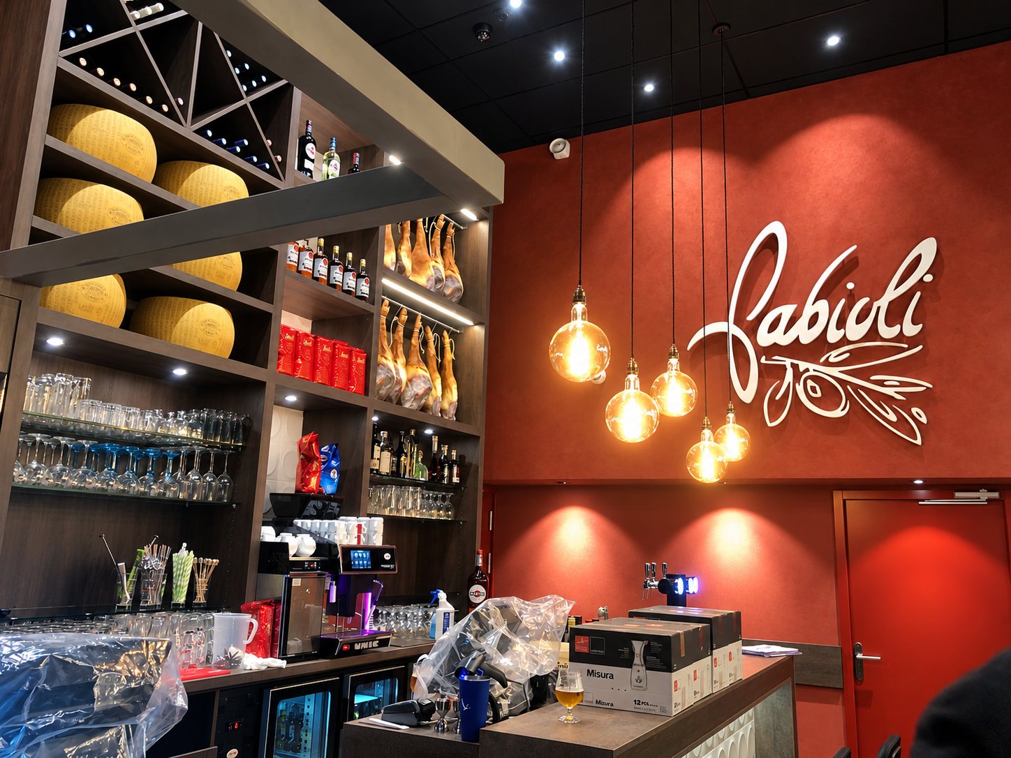 Bar rustique avec grandes meules de fromage et charcuterie sur des étagères, sous des suspensions à la lumière chaude.