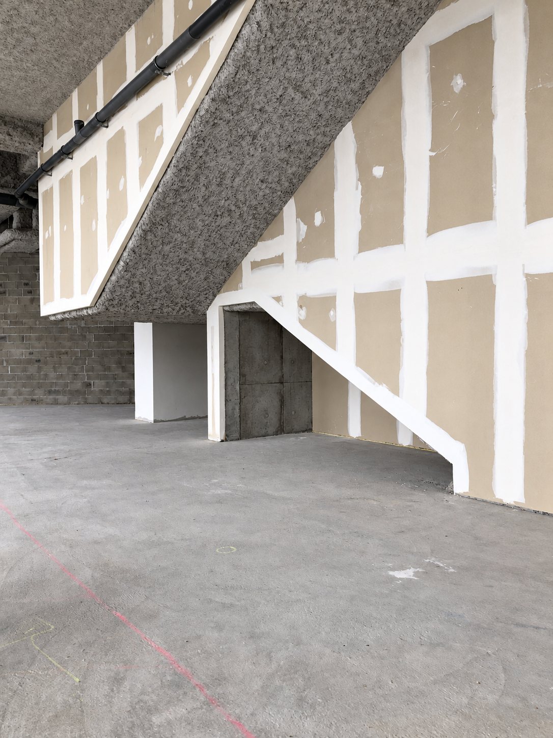 Chantier intérieur montrant des cloisons en plaques de plâtre aux joints enduits de blanc, sous un escalier en béton brut, avec un sol en béton gris.