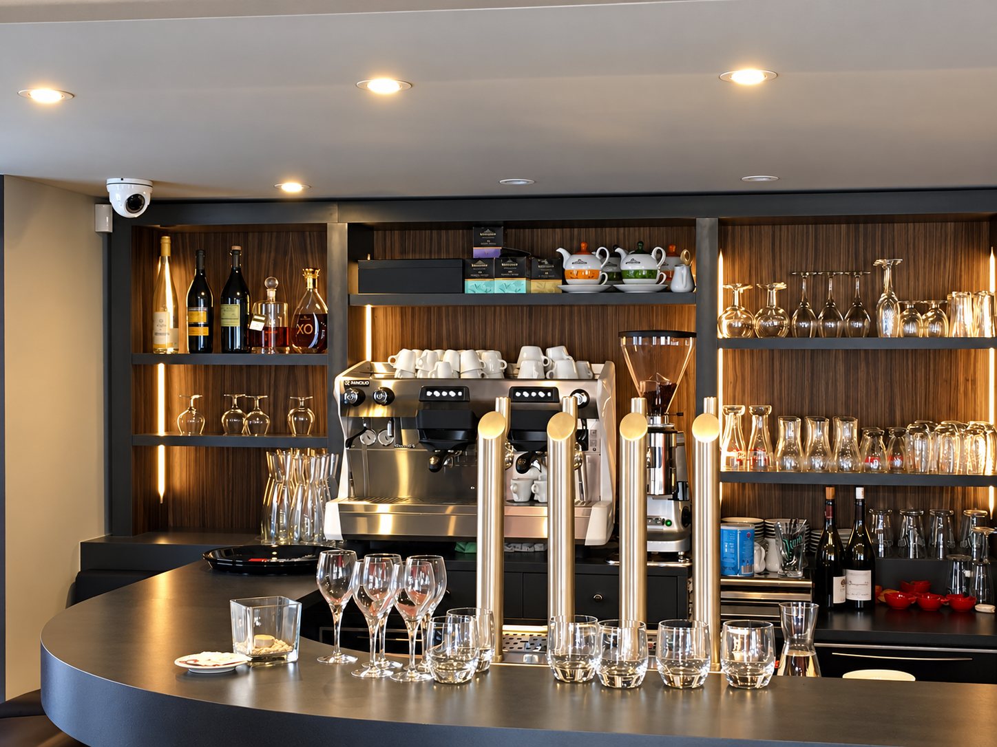 Un comptoir de bar arrondi en bois sombre avec quatre tireuses dorées, une machine à café, des tasses, des verres et des bouteilles sur les étagères.