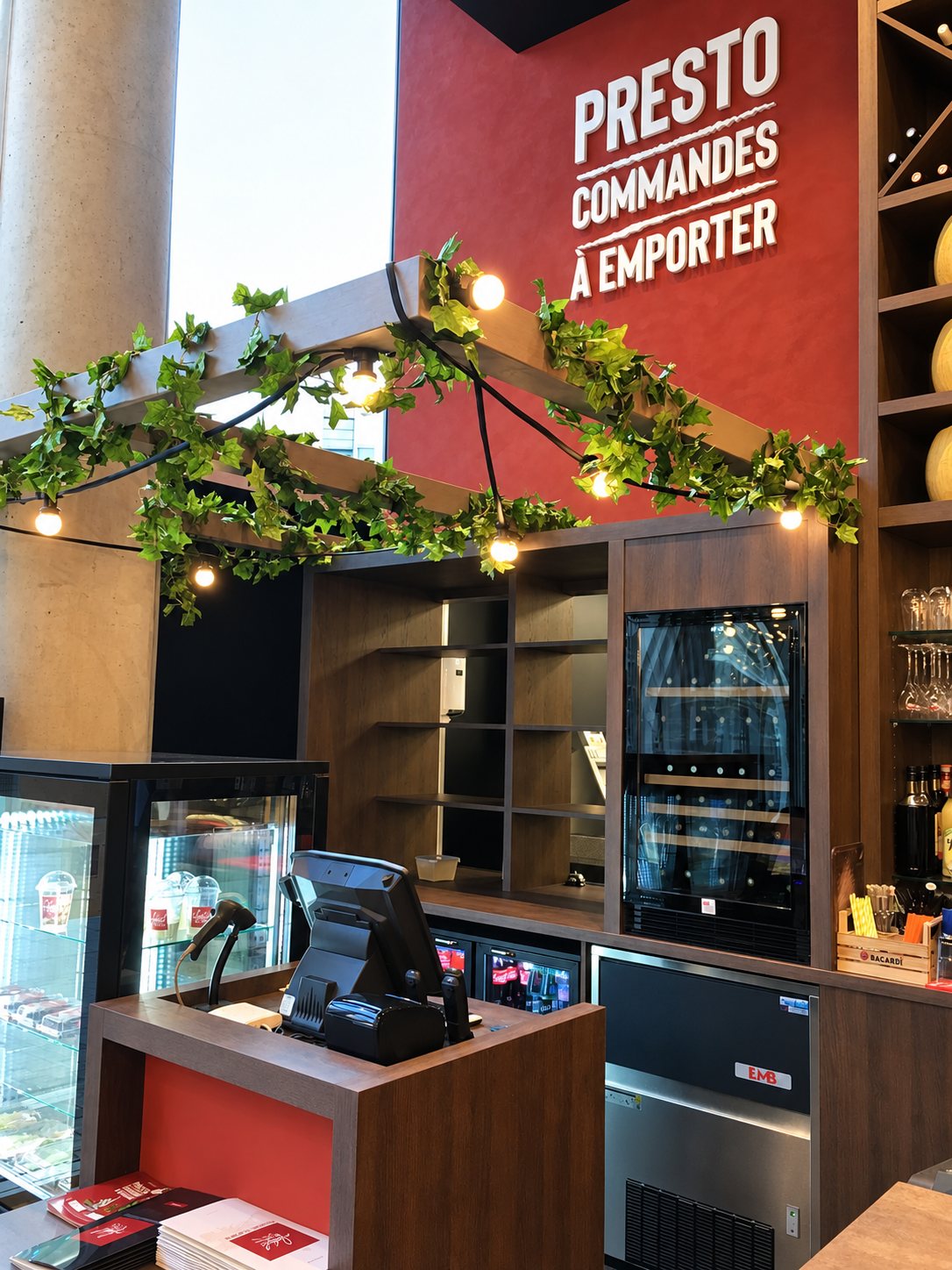 Comptoir de vente à emporter avec étagères en bois, petite cave à vin et élément végétal en vigne éclairé.