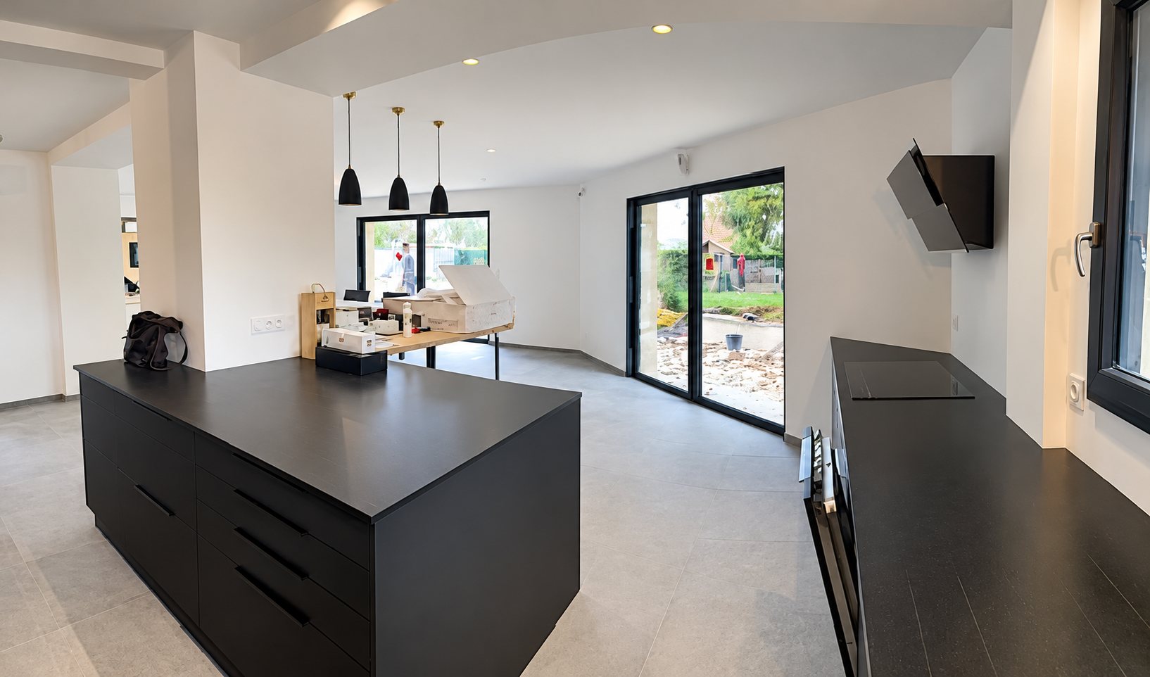 Vaste cuisine ouverte aux meubles noirs mats, avec un îlot central, une hotte inclinée et de grandes baies vitrées donnant sur le jardin.