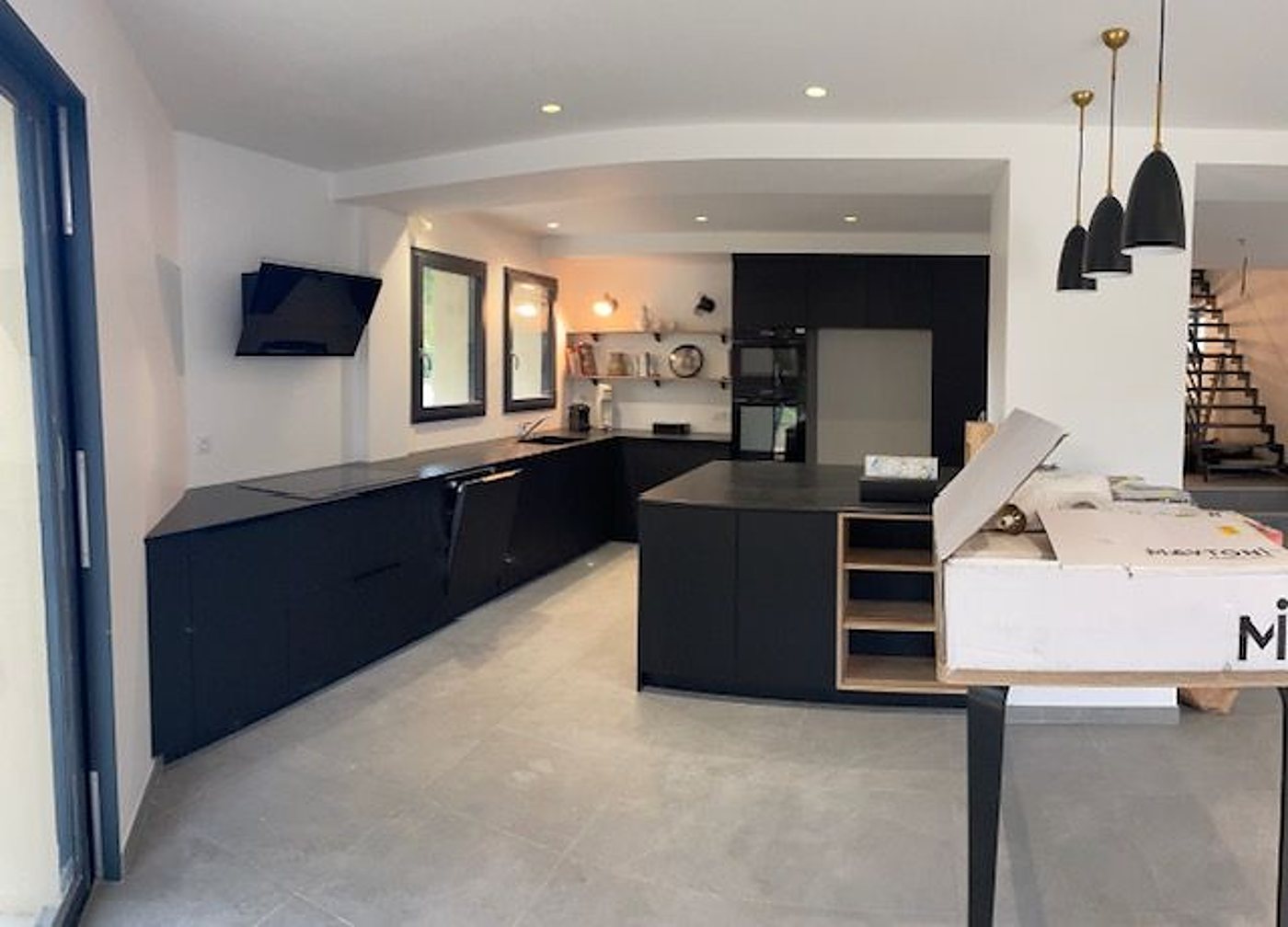 Cuisine contemporaine aux meubles noirs mats avec îlot central, suspensions noires et grand carrelage gris, ouverte sur un escalier.