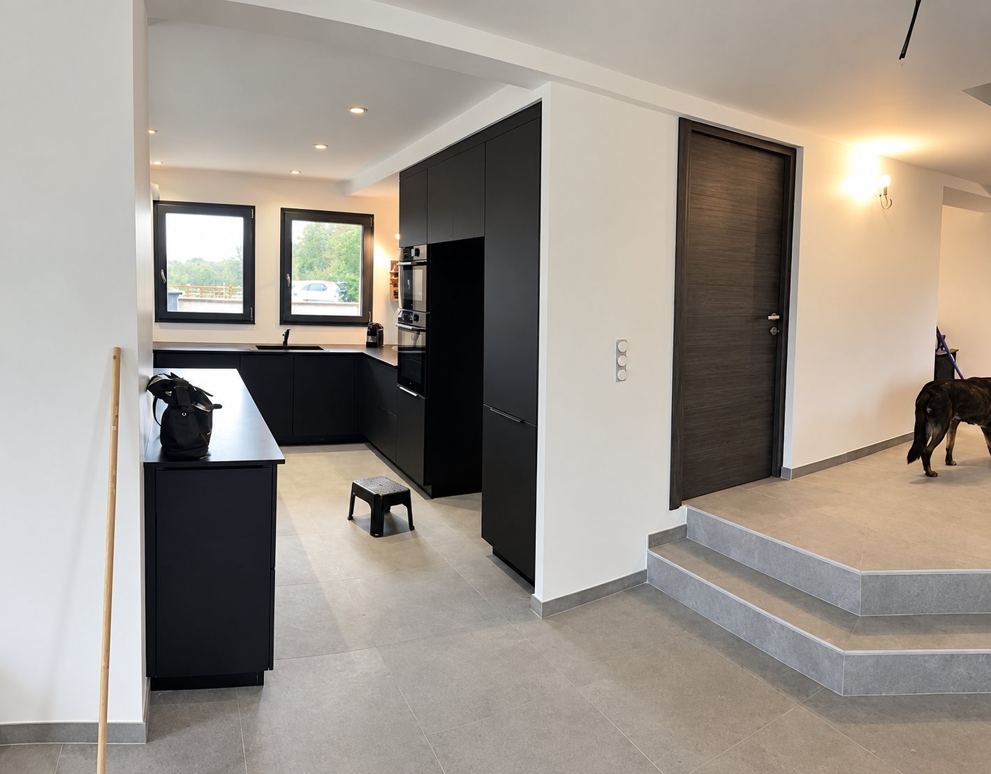 Une cuisine moderne aux meubles noirs mats avec îlot central, fenêtres noires et sol en pierre grise, à côté d'une porte en bois sombre et de deux marches.