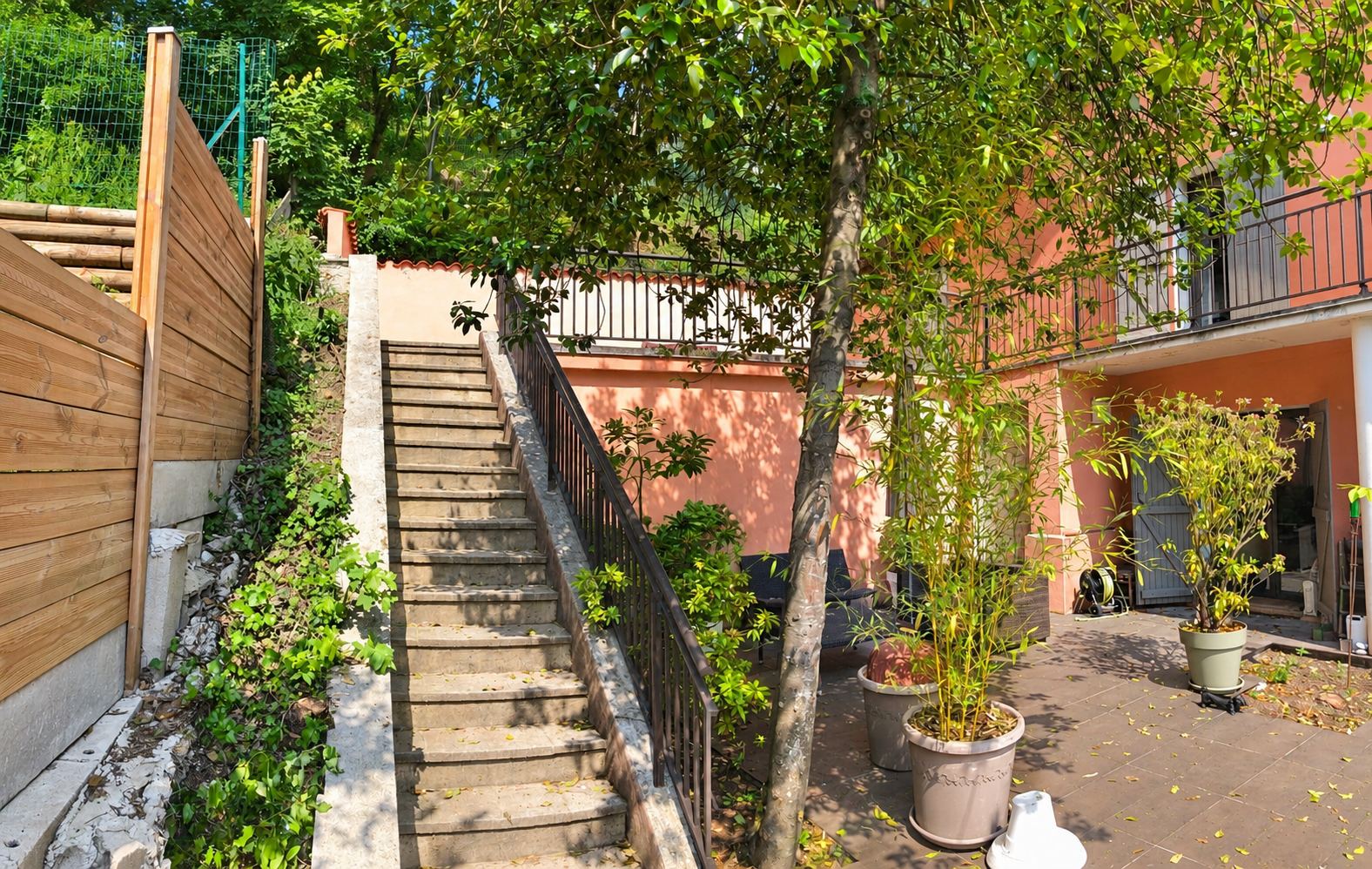 Un escalier en pierre avec rampe en fer forgé descend vers une terrasse dallée ornée de pots de plantes, à côté d'une clôture en bois et d'une maison rose.