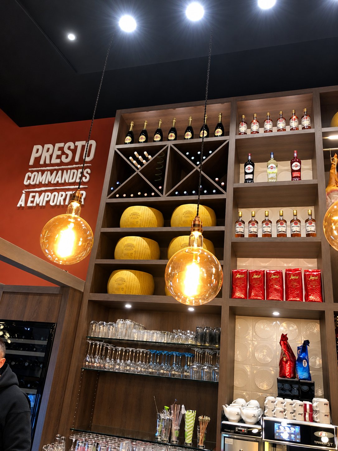 Étagère de restaurant avec bouteilles de vin, meules de fromage, verres et sacs décoratifs rouges.