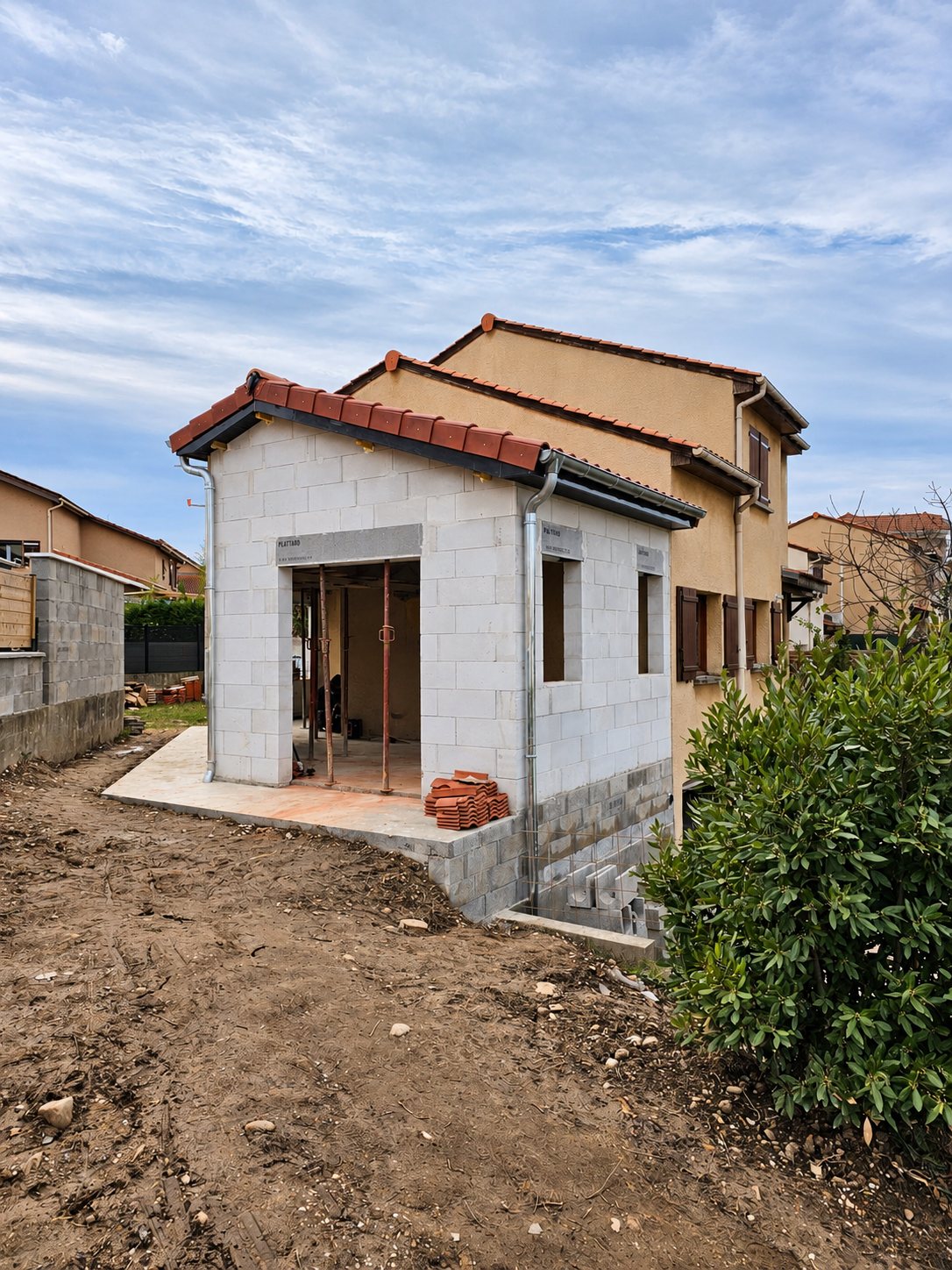 Extension récente en béton clair accolée à une maison beige à étage.