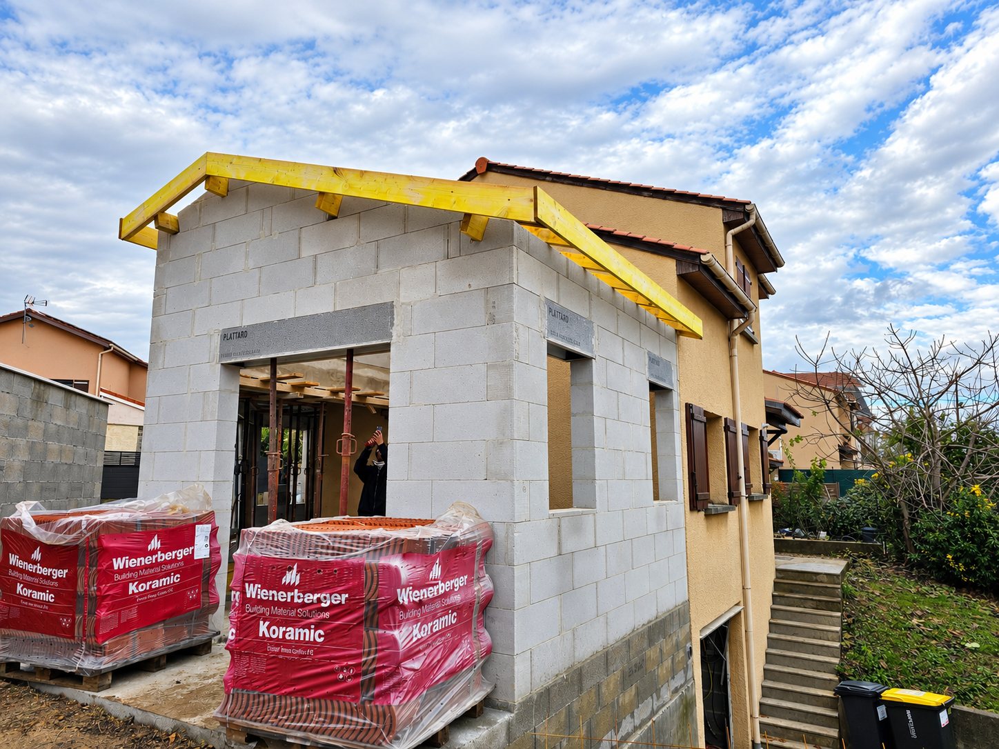Extension en parpaings inachevée à côté d'une maison beige, avec des palettes de briques filmées à proximité.