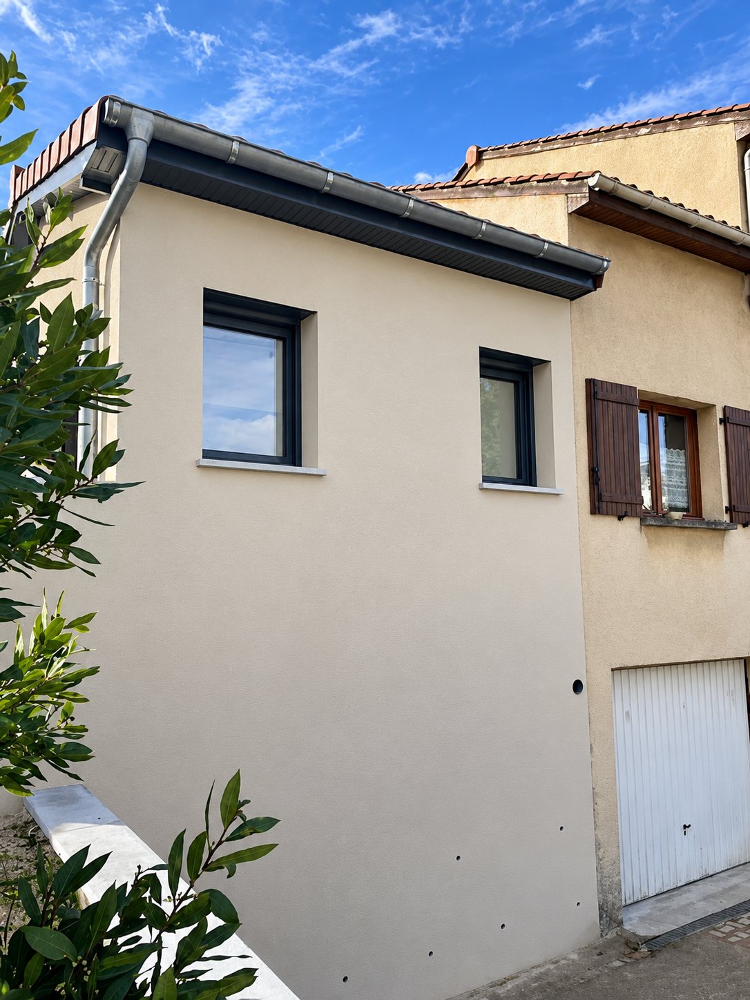 Façade crème avec deux fenêtres et une gouttière, à côté d'une maison plus ancienne avec garage.