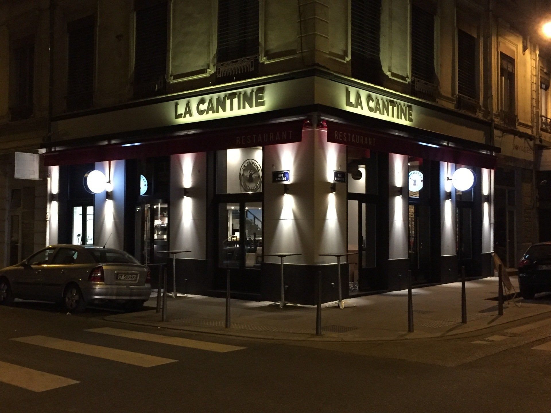 Façade d'angle illuminée de nuit du restaurant La Cantine, avec ses tables hautes en terrasse et une voiture garée devant.