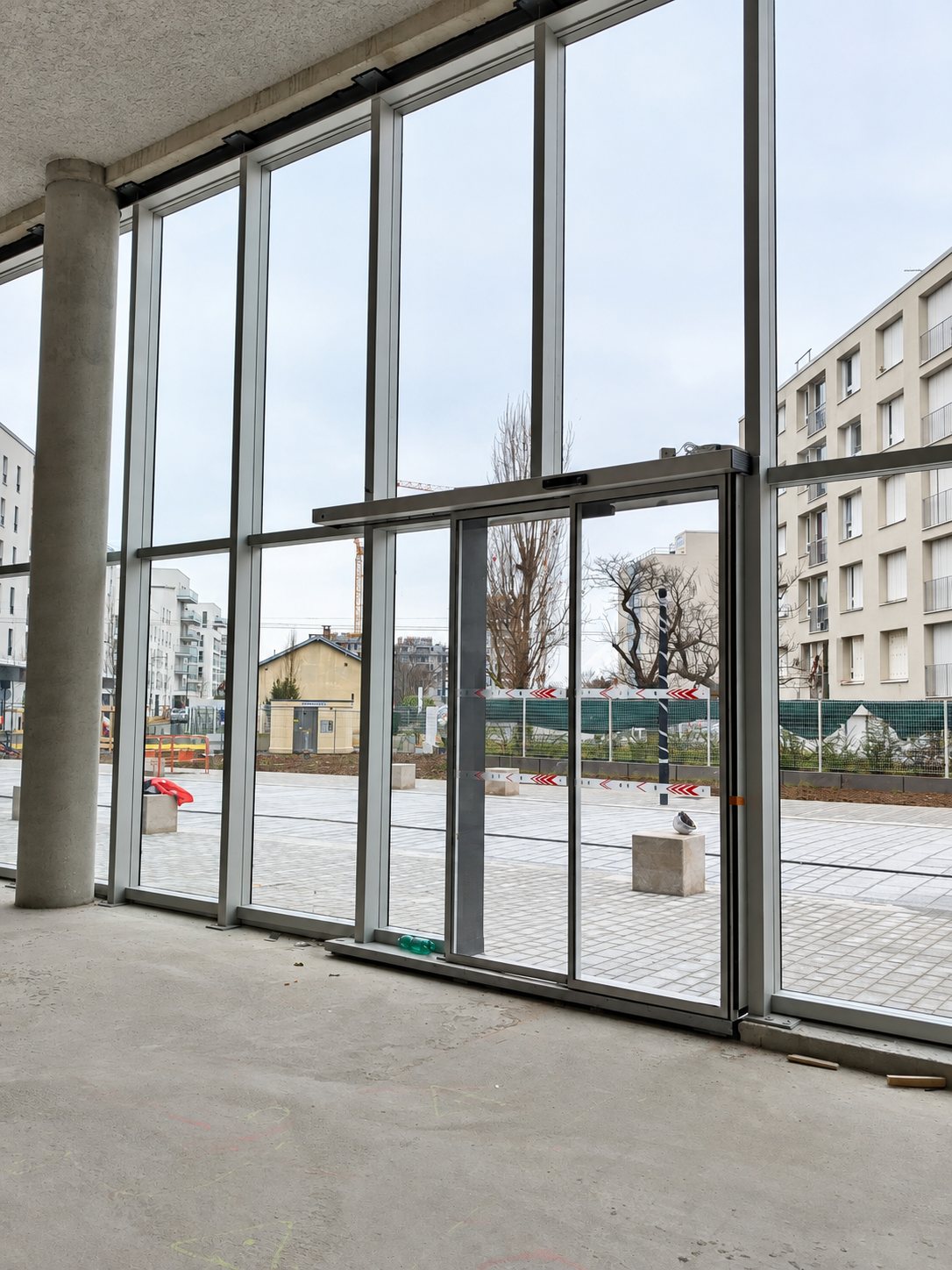 Vue depuis l'intérieur d'un local en chantier sur une grande façade vitrée équipée d'une porte automatique coulissante donnant sur une place pavée et des immeubles.