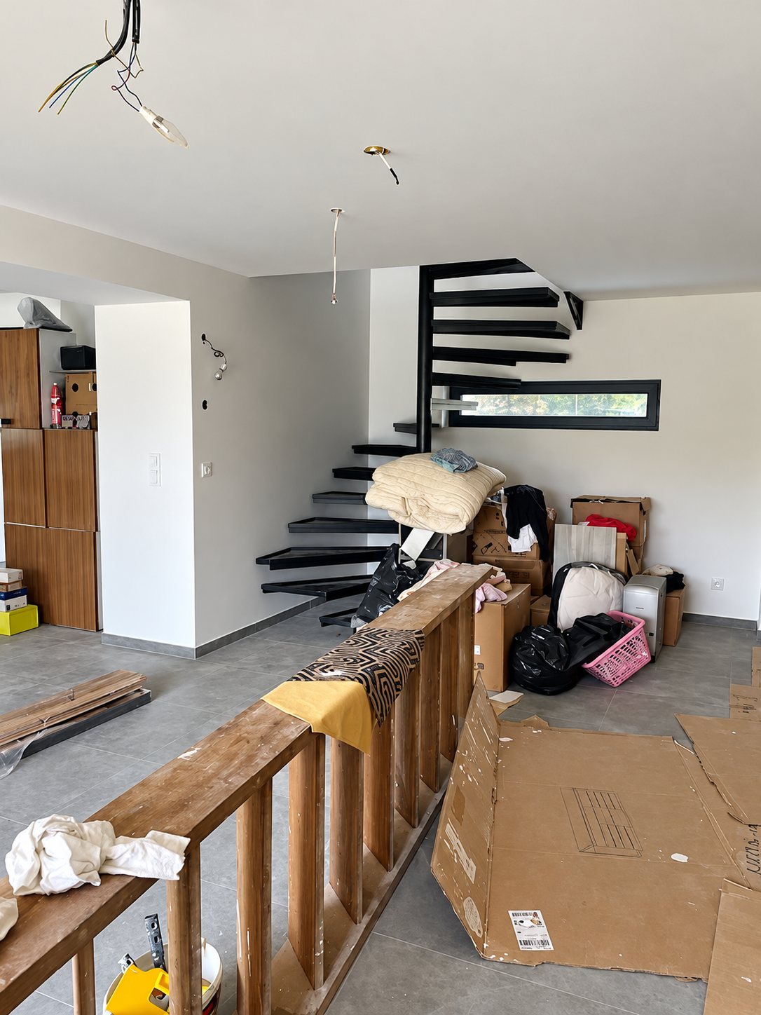 Pièce de vie en travaux avec un escalier noir à marches suspendues, une cloison en bois en construction et des cartons posés sur le carrelage gris.