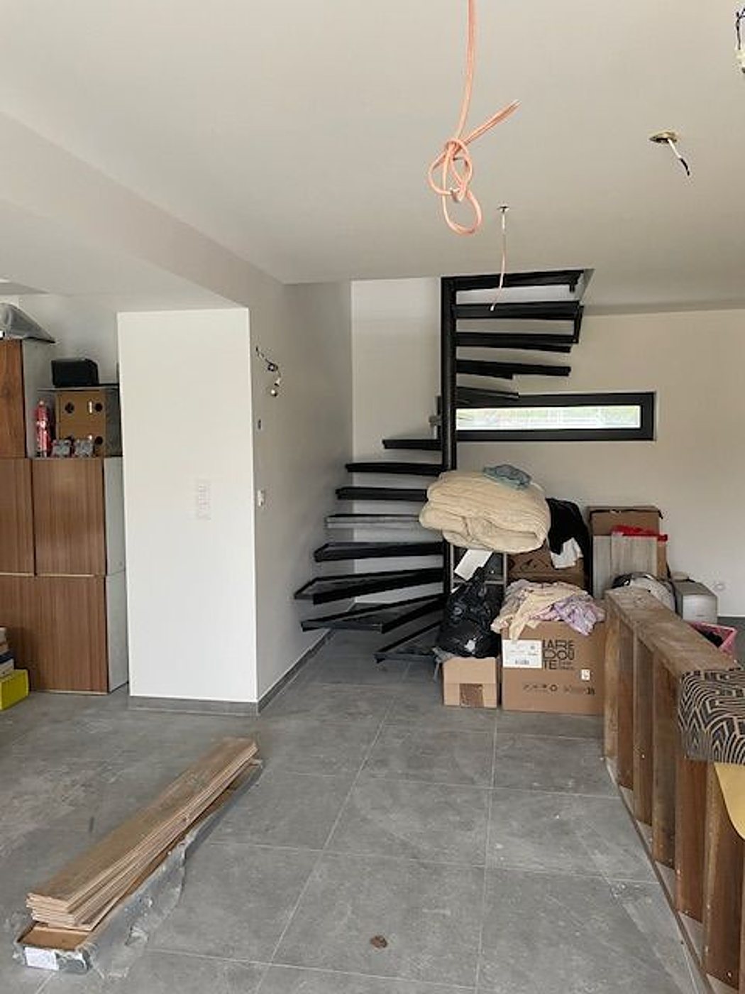 Pièce en rénovation avec un escalier en colimaçon noir, des cartons et des couvertures empilés, des lames de parquet posées au sol carrelé gris.