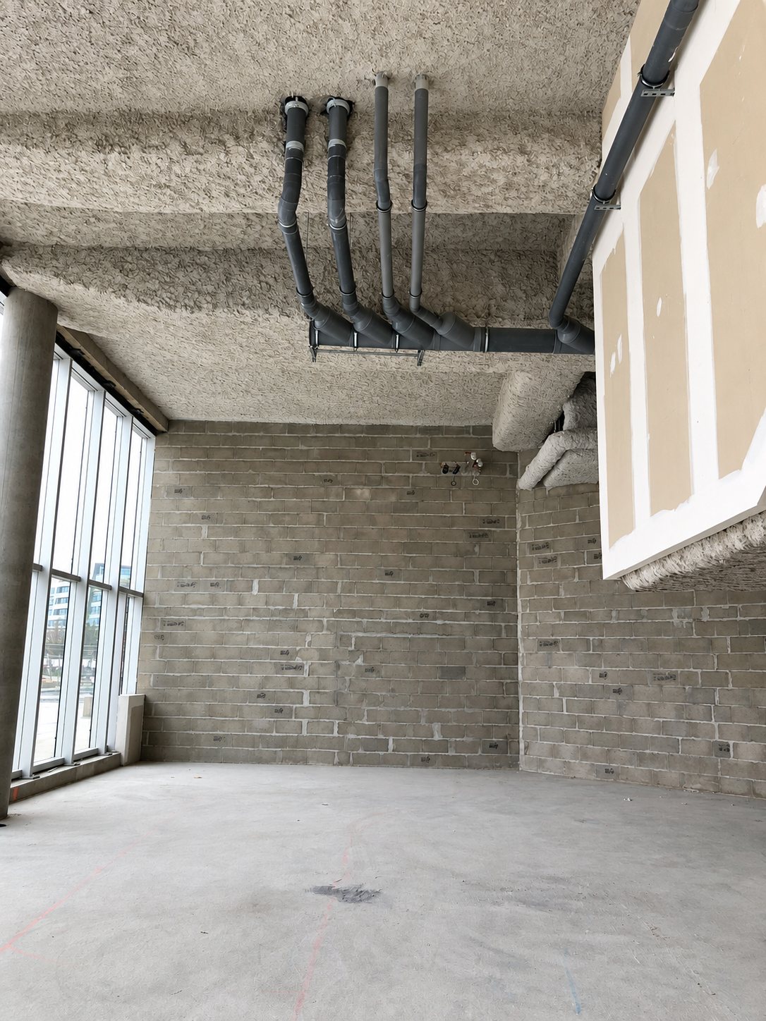 Pièce en chantier avec murs en parpaings, dalle béton brute, grande baie vitrée et canalisations grises apparentes au plafond isolé.