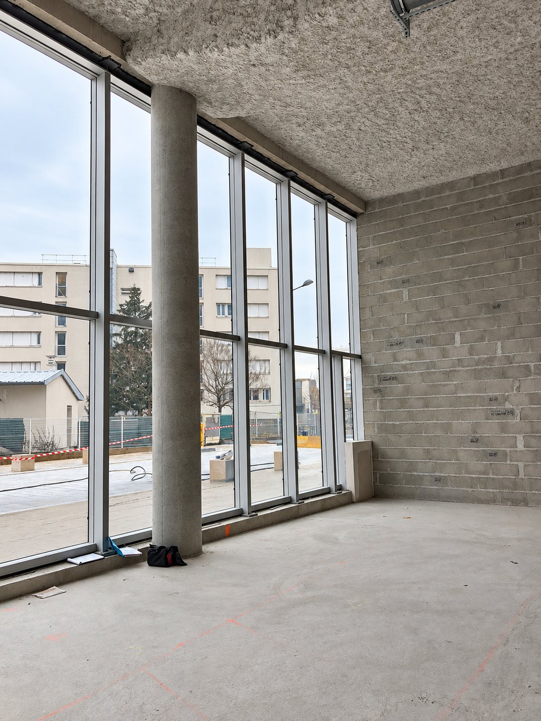 Intérieur d'un local en chantier avec une grande façade vitrée, un pilier en béton, un mur en parpaings bruts et un plafond isolant floqué.