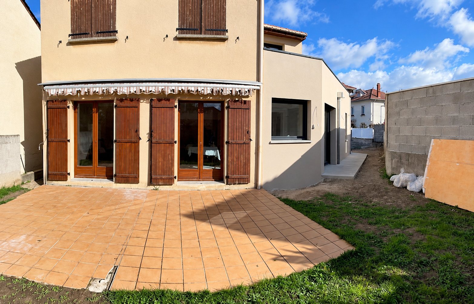 Maison beige avec portes-fenêtres, volets en bois et terrasse carrelée, à côté d'une cour non goudronnée.
