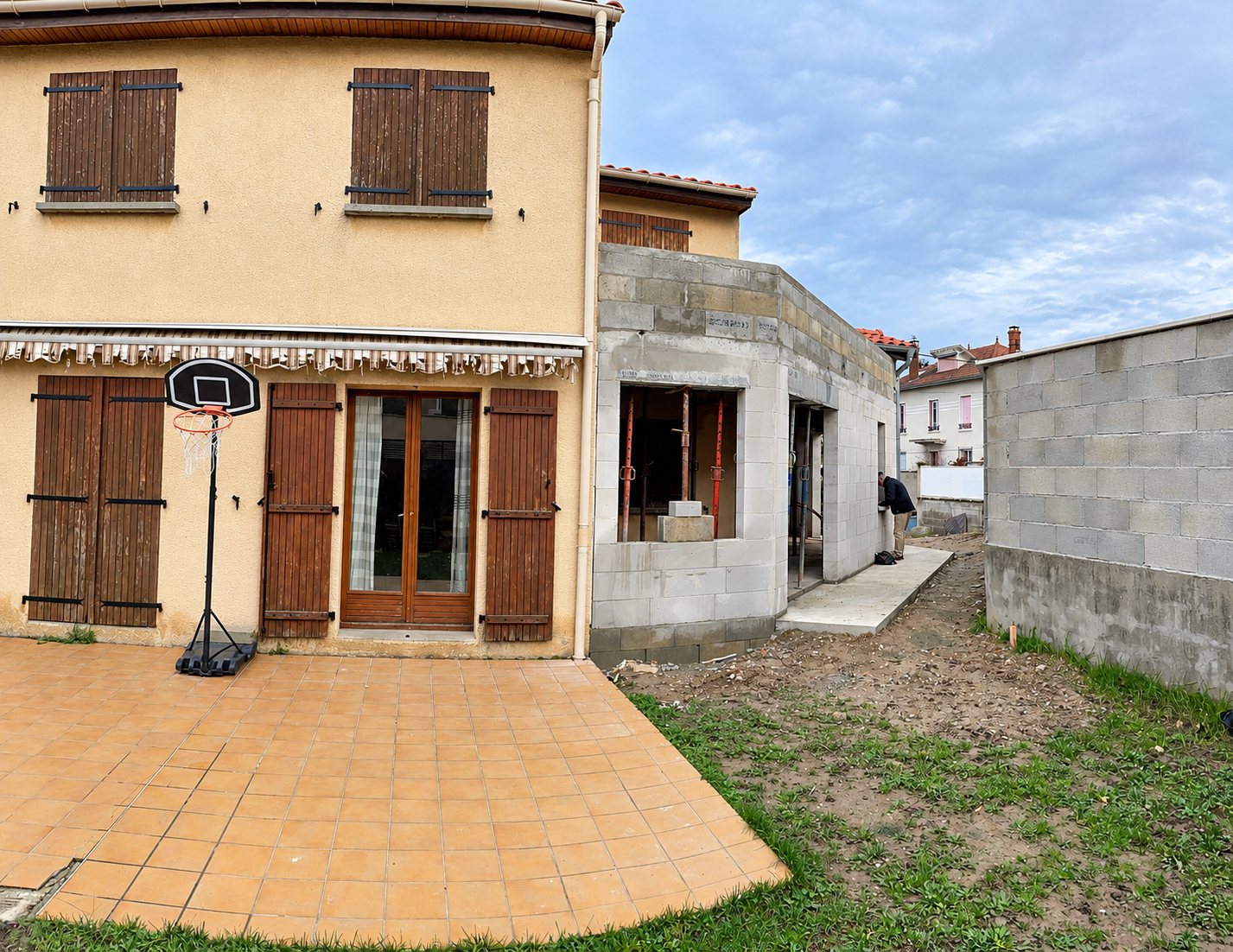 Maison beige à étage avec volets marron, à côté d'une extension en construction.
