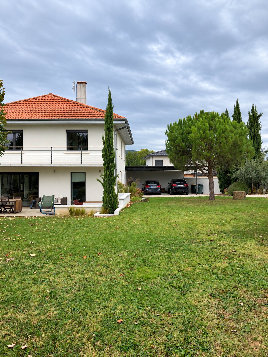 Maison blanche à étage avec toit en tuiles orange, terrasse, pelouse, pin parasol et deux voitures garées sous un carport.