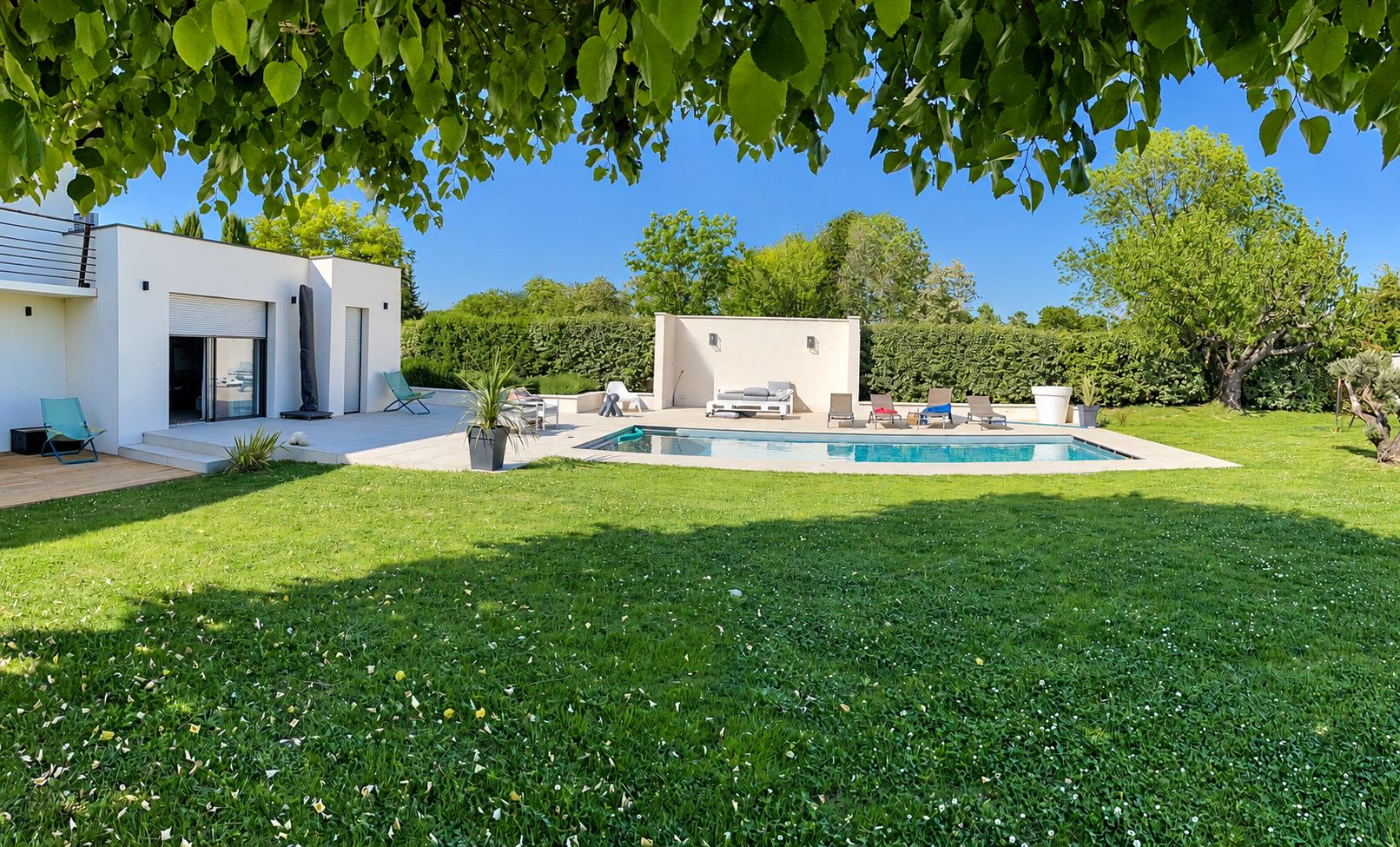 Une maison contemporaine blanche avec une terrasse et une piscine rectangulaire entourées d'une grande pelouse et de haies sous un ciel bleu.