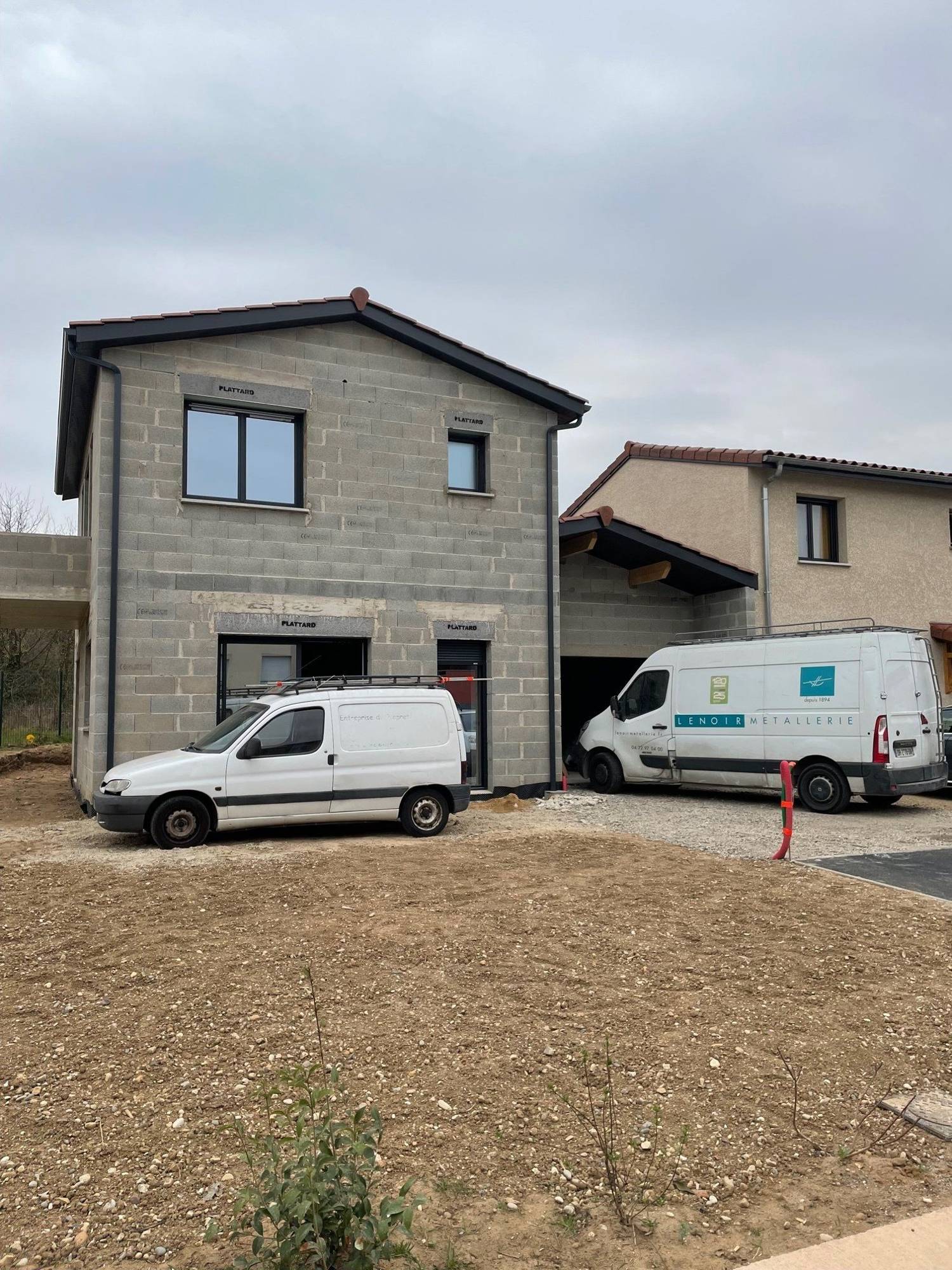 Une maison en parpaings en cours de construction, avec des menuiseries noires posées, devant laquelle sont garées deux camionnettes blanches dont une de la métallerie Lenoir.