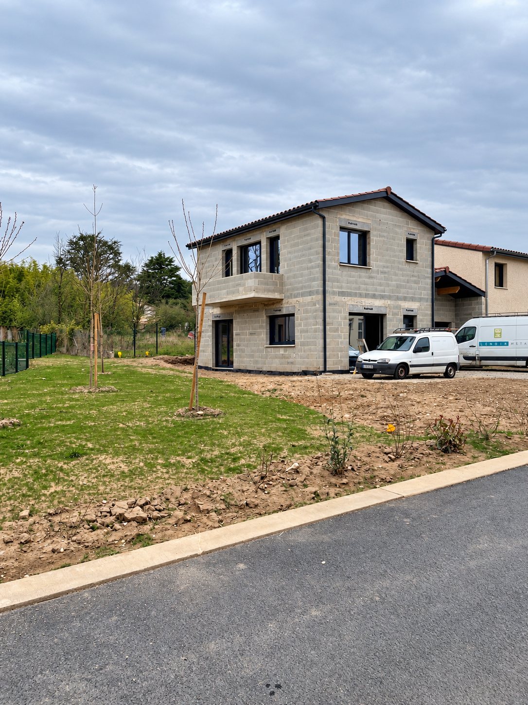 Une maison en parpaings apparents avec balcon, en cours de construction, devant laquelle sont garées deux camionnettes blanches, sur un terrain en cours d'aménagement.