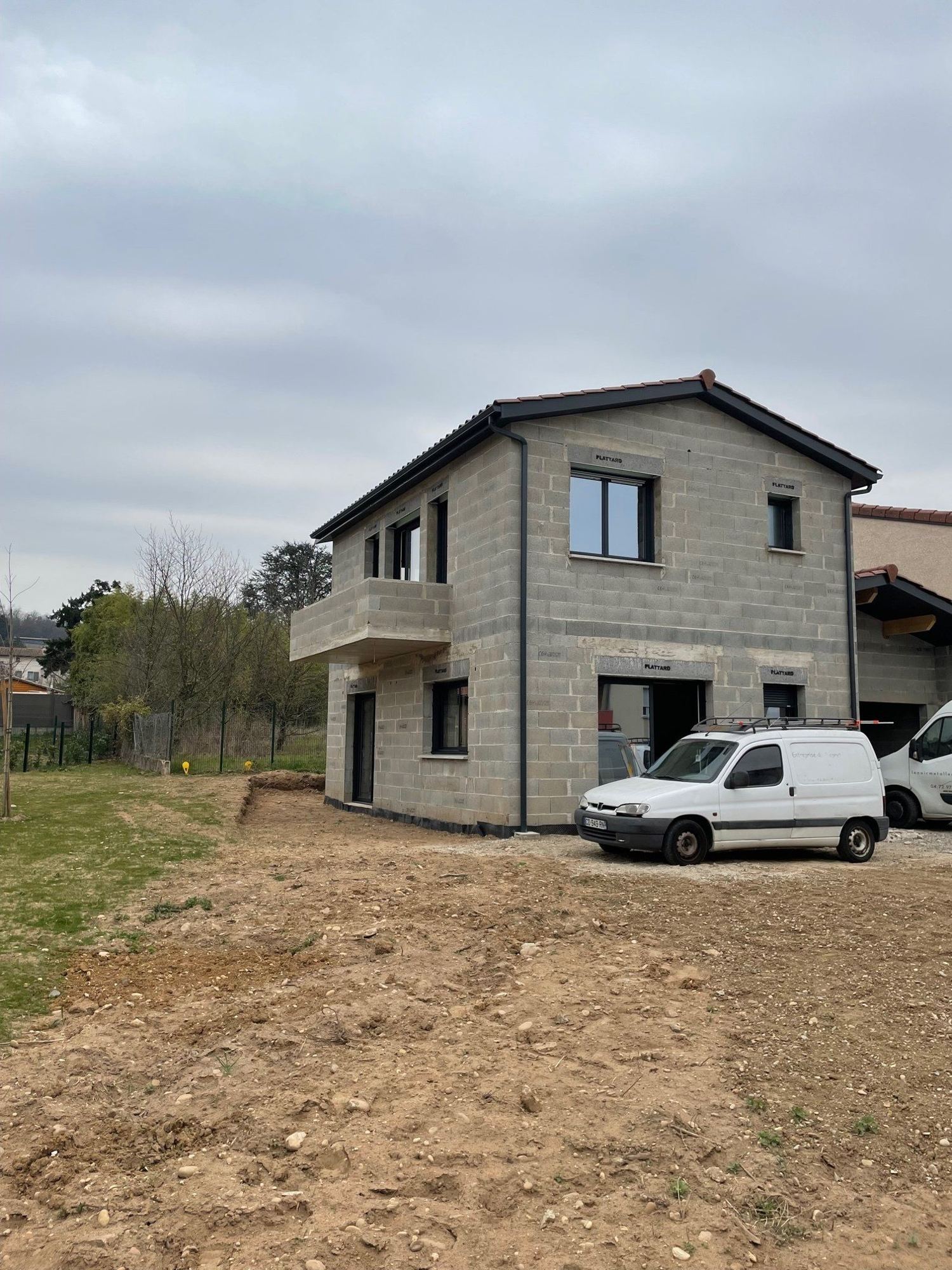 Une maison à étage en blocs de béton bruts, dotée de menuiseries sombres et d'un toit en tuiles, devant laquelle est garée une camionnette blanche sur un terrain de terre.