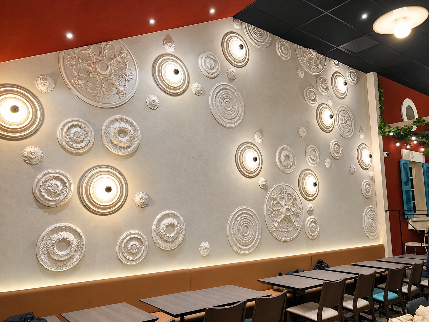 Un grand mur gris orné de rosaces en plâtre et de appliques lumineuses circulaires, au-dessus d'une banquette et de tables en bois dans une salle de restaurant.