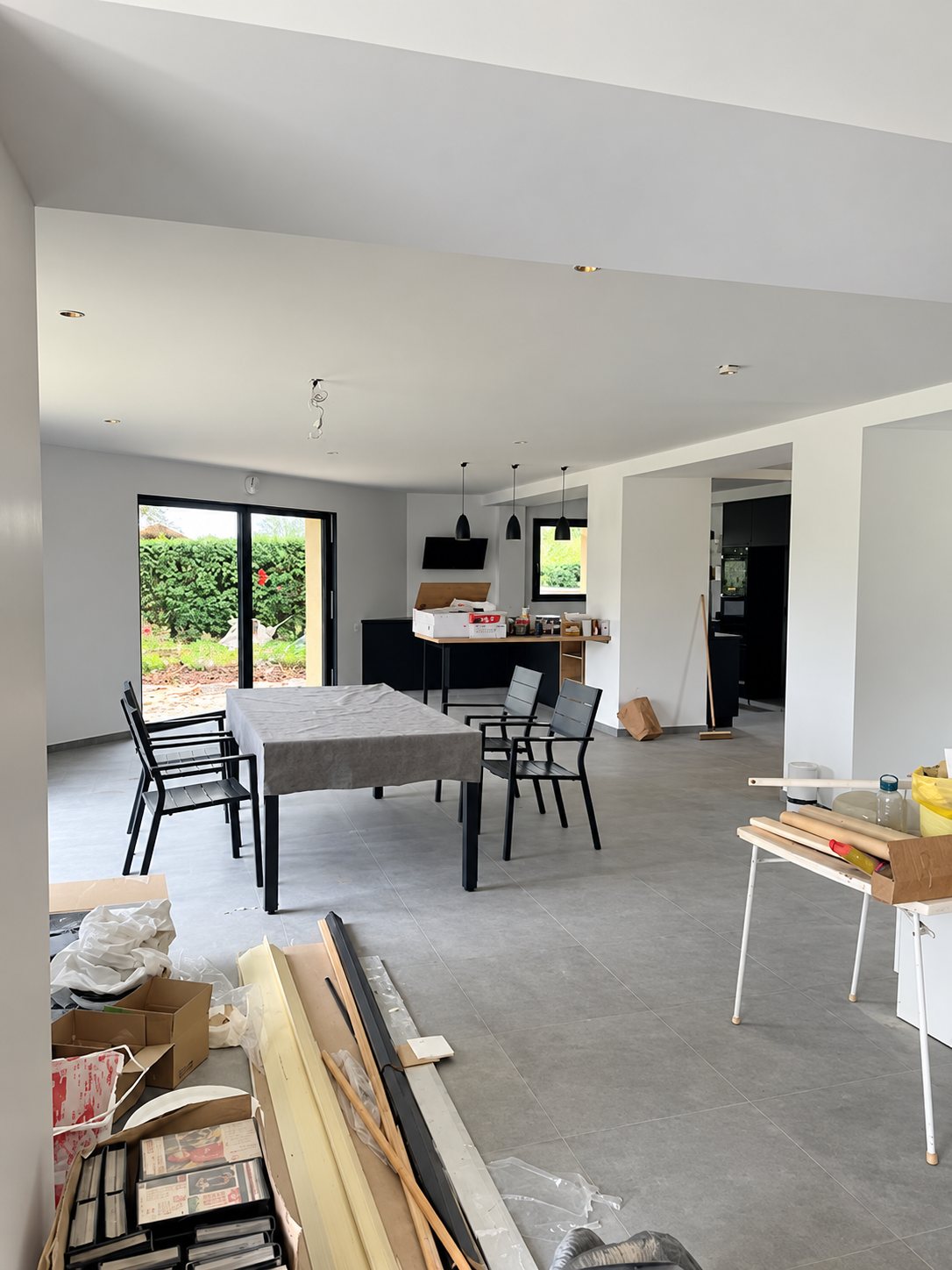 Grande pièce à vivre lumineuse en cours de finition, avec table grise, chaises noires, baie vitrée et matériaux de chantier au premier plan.