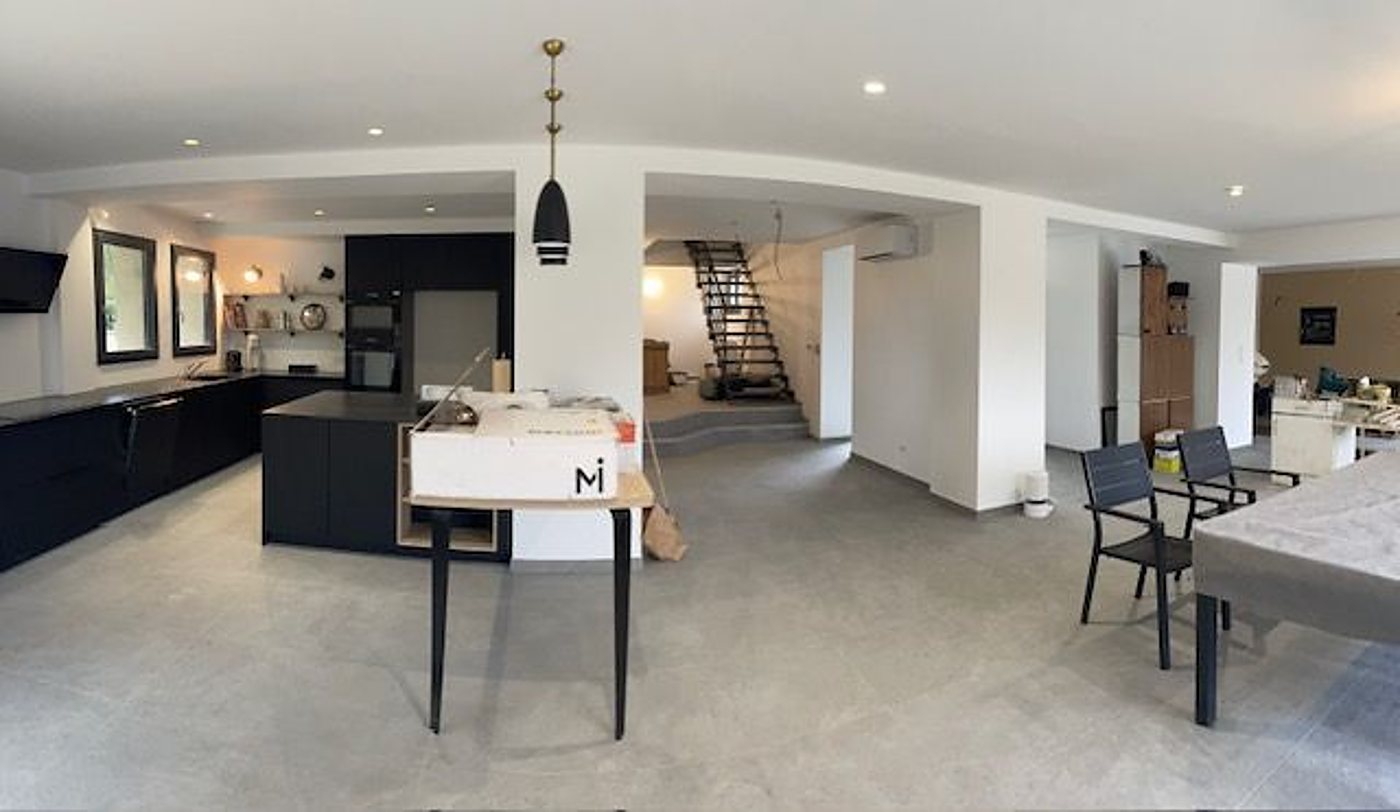 Vue panoramique d'un grand espace ouvert au sol en carrelage gris, avec une cuisine noire à îlot, un escalier métallique et une table avec des chaises au fond.