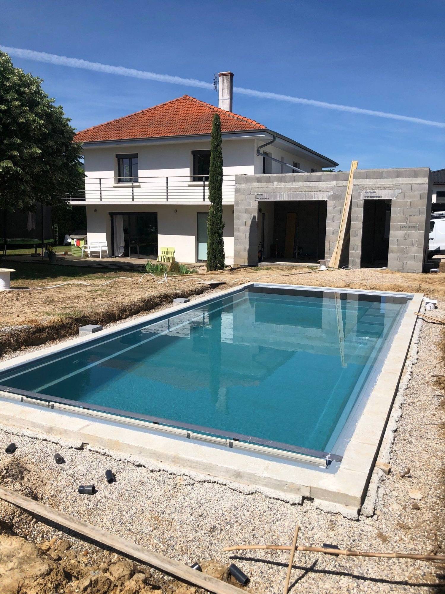 Une piscine rectangulaire à l'eau turquoise, entourée de gravier et de terre, est en cours d'aménagement devant une maison blanche à toit de tuiles rouges et son extension en parpaings.