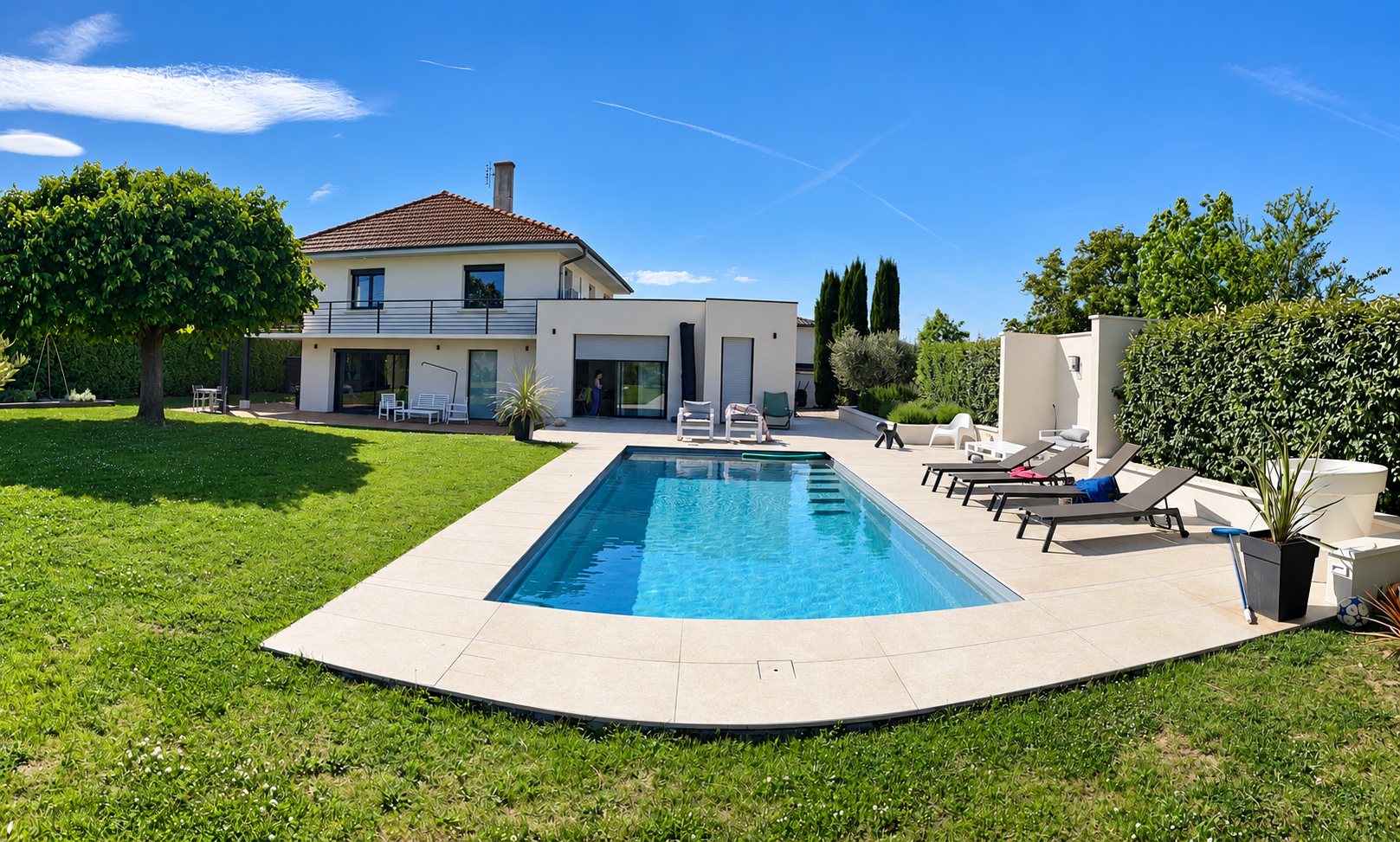 Une piscine rectangulaire entourée d'une terrasse claire et de transats, avec une maison à étage et une pelouse verte en arrière-plan.