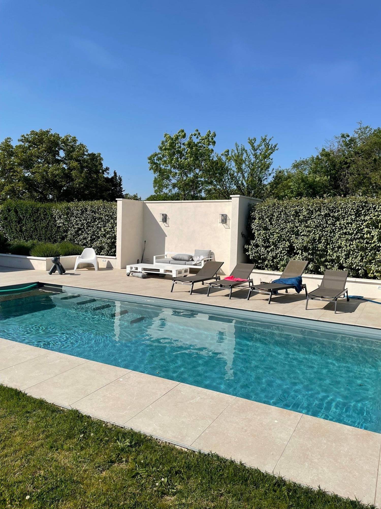 Une piscine rectangulaire à l'eau turquoise entourée d'une plage en dalles beiges, de chaises longues, d'un salon de jardin et de haies vertes sous un ciel bleu.
