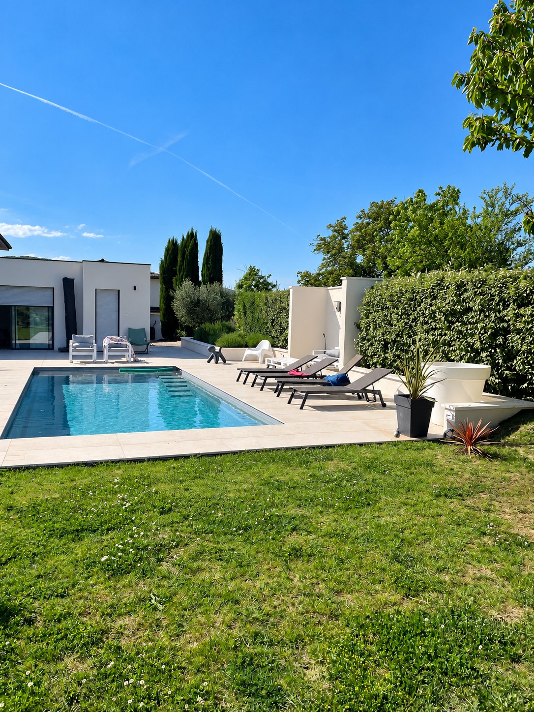 Une piscine rectangulaire à l'eau turquoise entourée d'une terrasse claire, de transats, d'une maison moderne, de cyprès et d'une pelouse verte sous un ciel bleu.