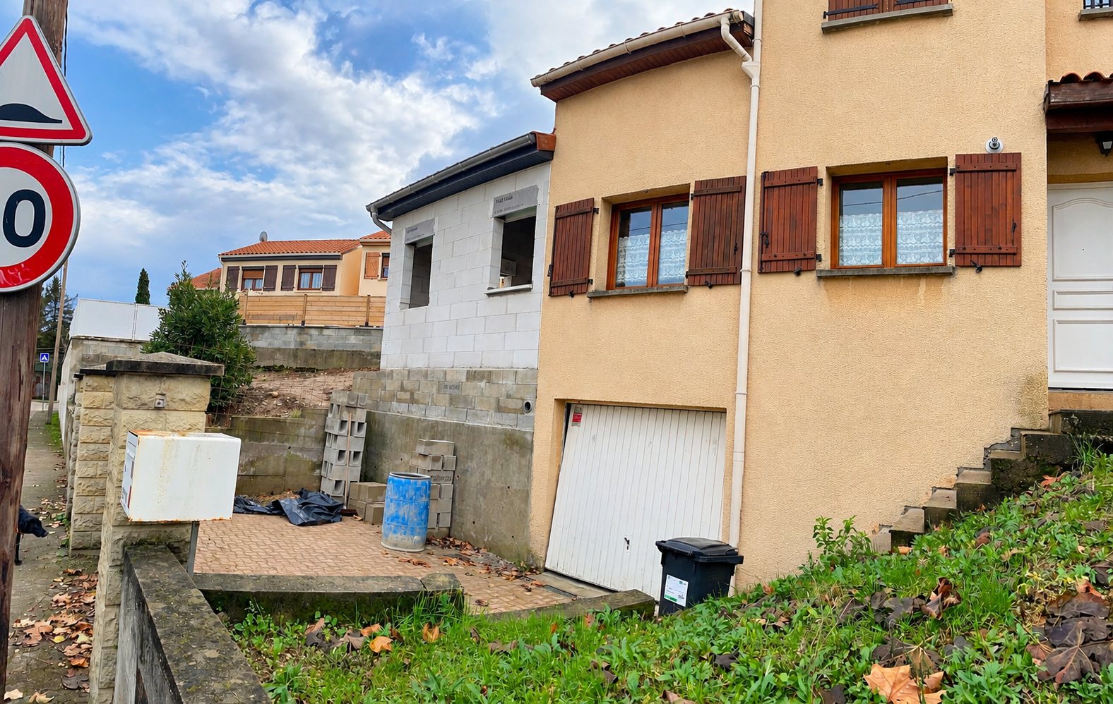 Vue depuis la rue d'une maison beige à étage avec volets marron et garage blanc, à côté d'un petit bâtiment blanc.