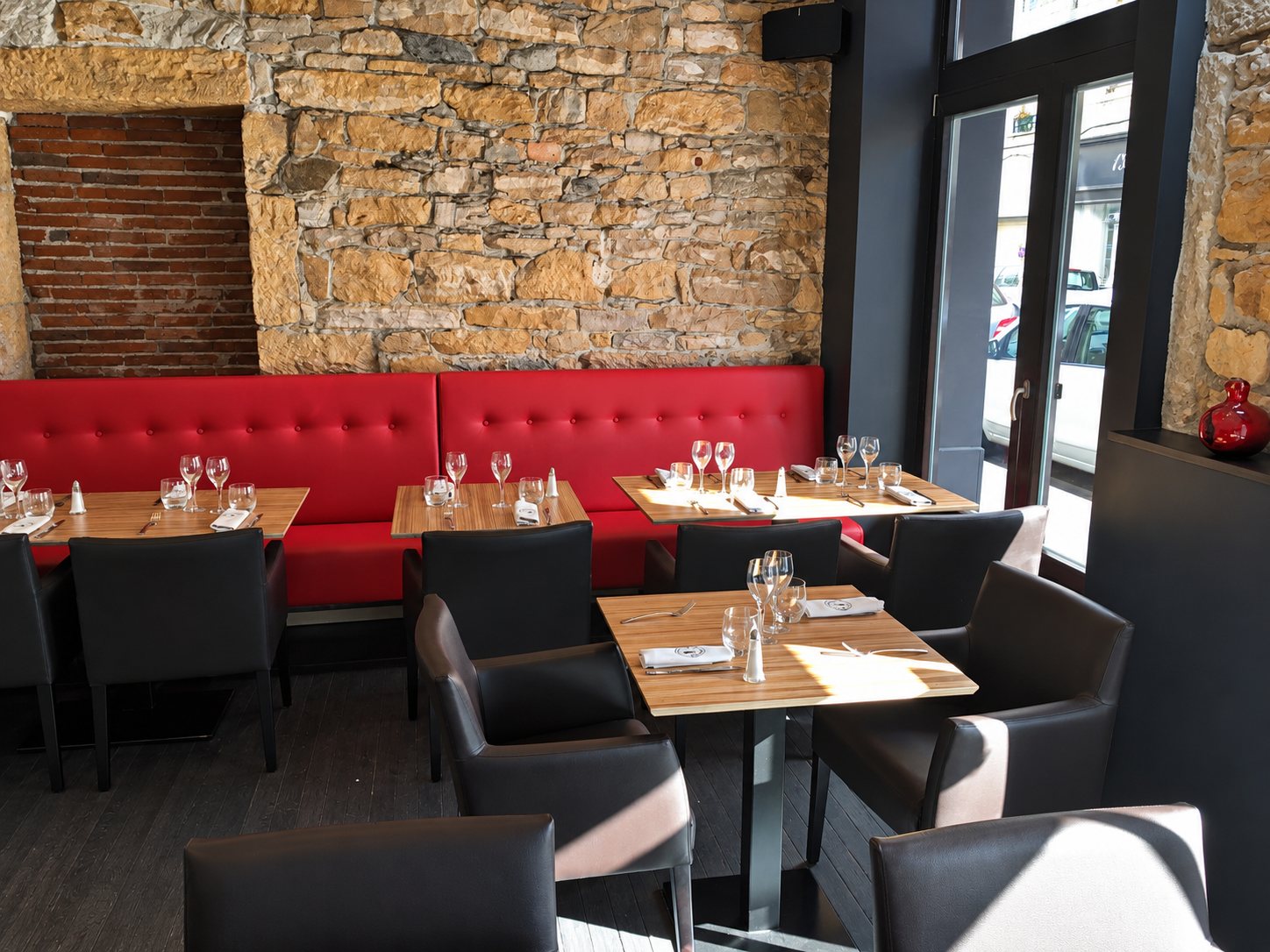 Intérieur d'un restaurant avec des tables en bois dressées, une banquette rouge capitonnée et un mur de pierres apparentes près d'une grande baie vitrée.