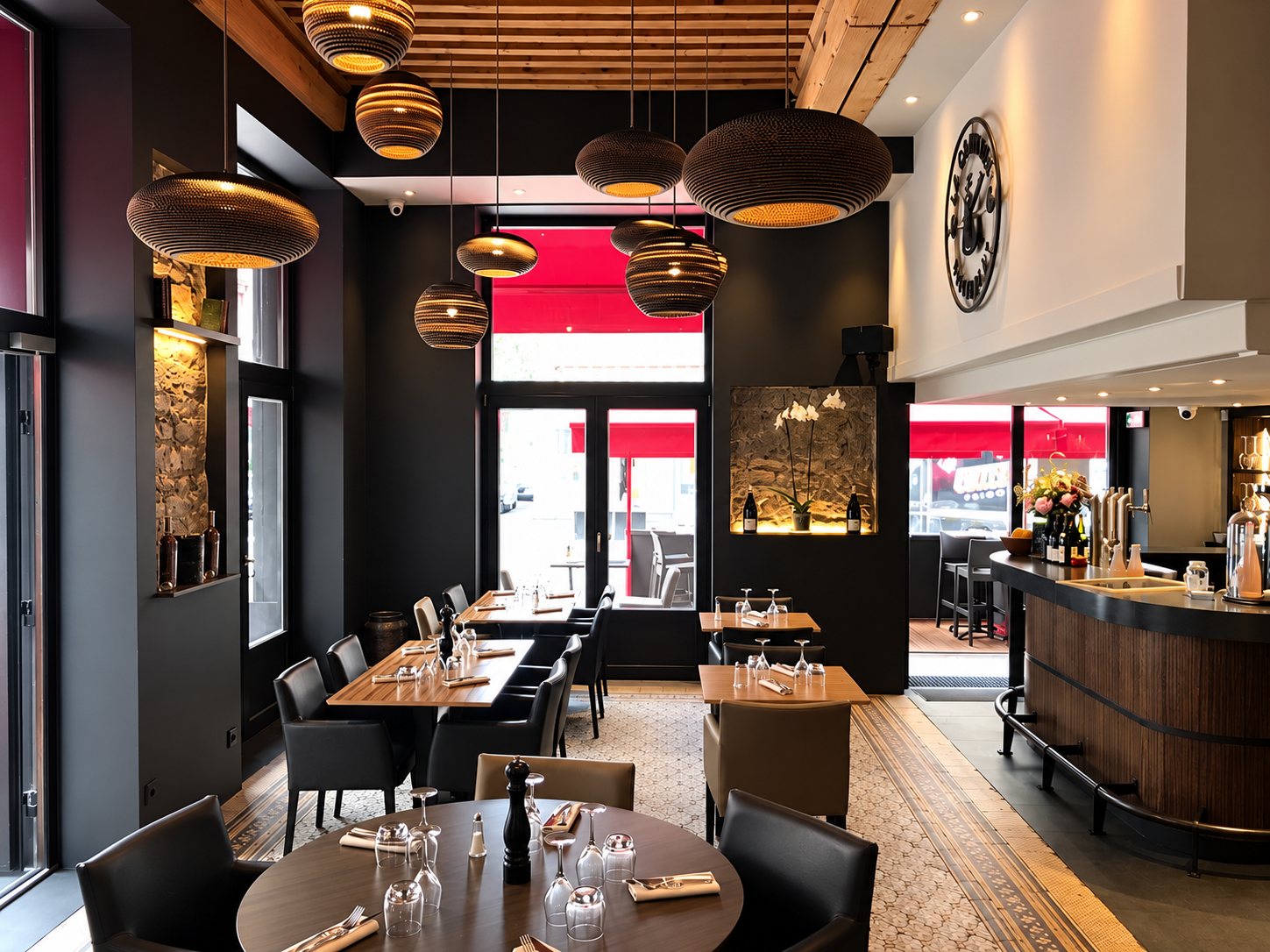 Intérieur d'un restaurant aux murs sombres avec tables dressées, suspensions en carton ondulé, pierre apparente et bar en bois incurvé.
