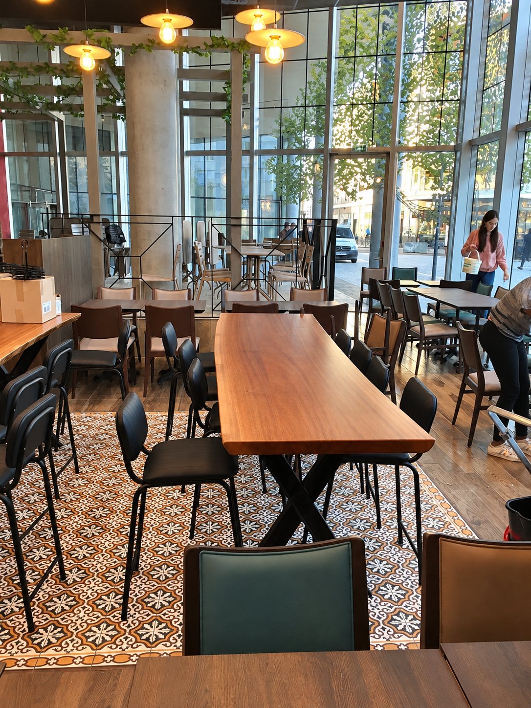 Intérieur d'un restaurant aux grandes baies vitrées, avec une longue table en bois, des chaises noires et un sol en carreaux de ciment décorés.