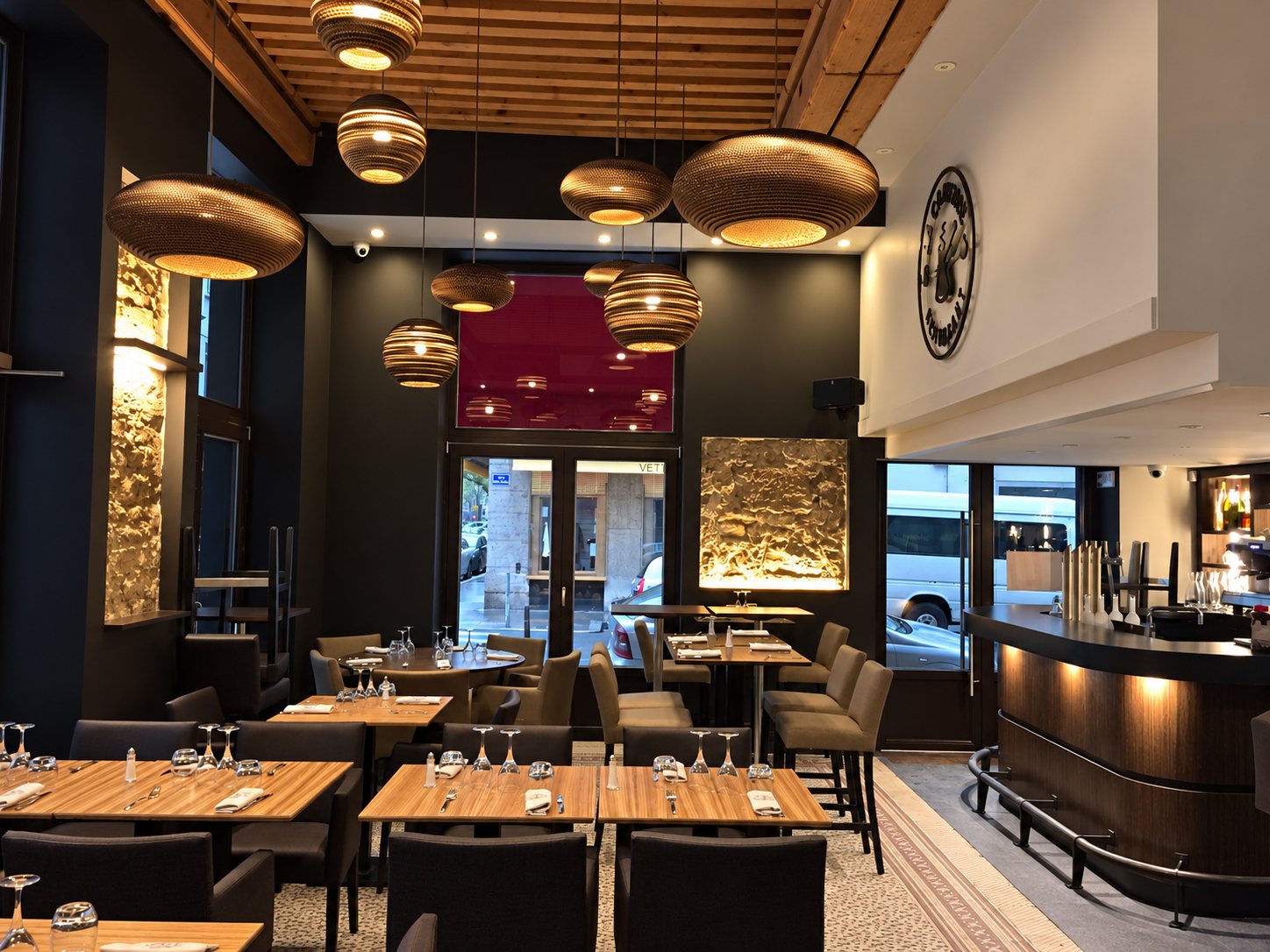 Intérieur d'un restaurant aux tons sombres avec tables en bois dressées, grandes suspensions en carton ondulé, murs en pierre éclairés et comptoir de bar courbe.
