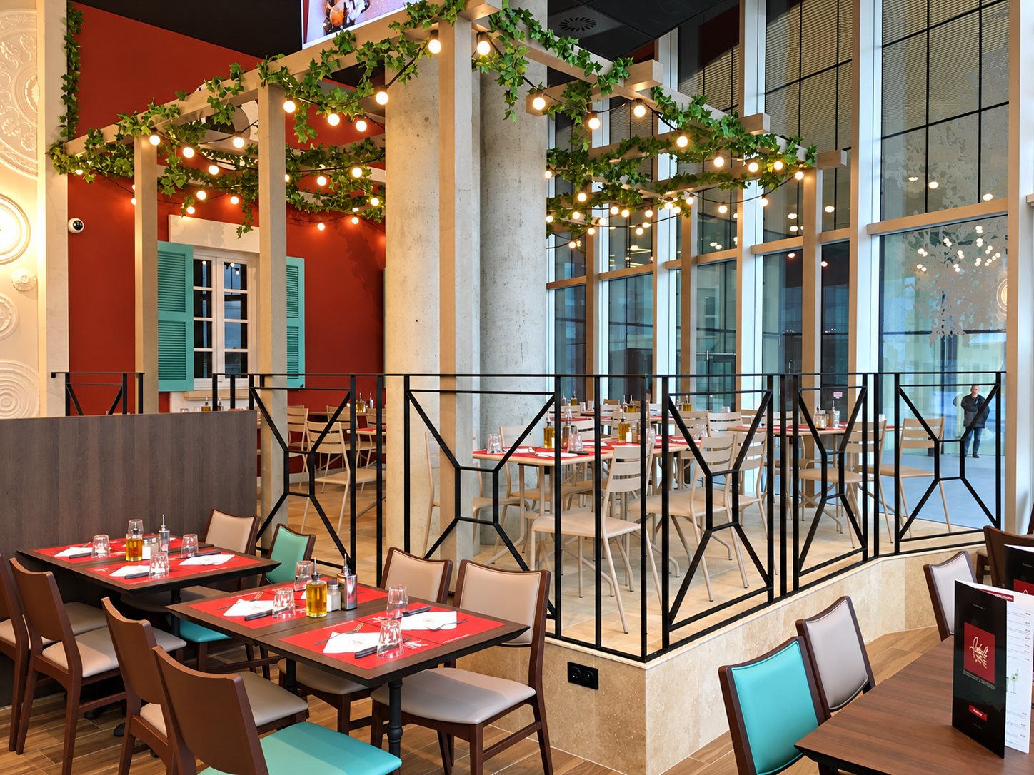 Intérieur d'un restaurant avec tables aux sets rouges, chaises en bois et turquoise, guirlandes lumineuses et grandes baies vitrées.