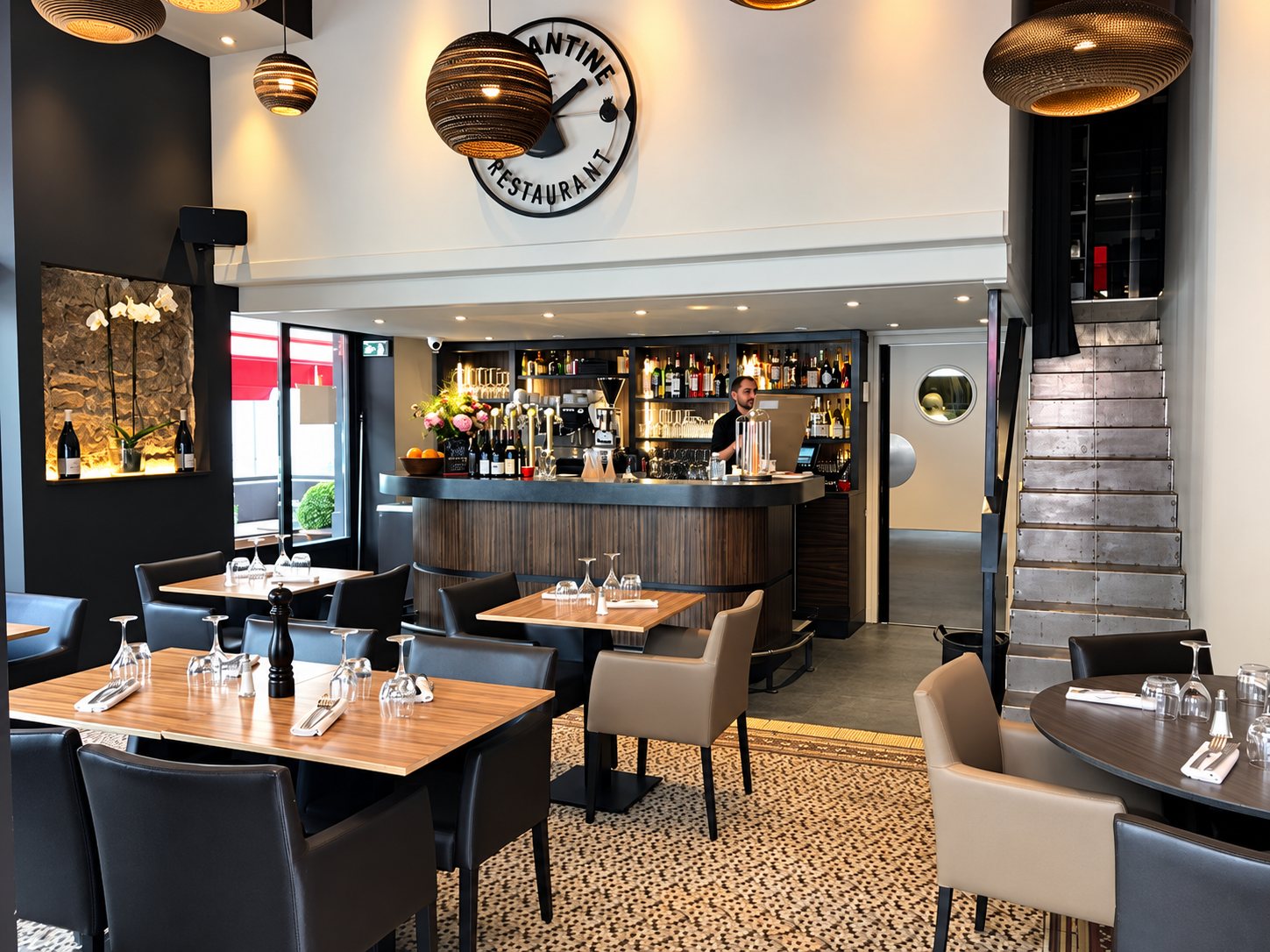 Intérieur d'un restaurant avec tables en bois dressées, fauteuils sombres et beiges, un bar en arrondi au fond et un escalier métallique à droite.