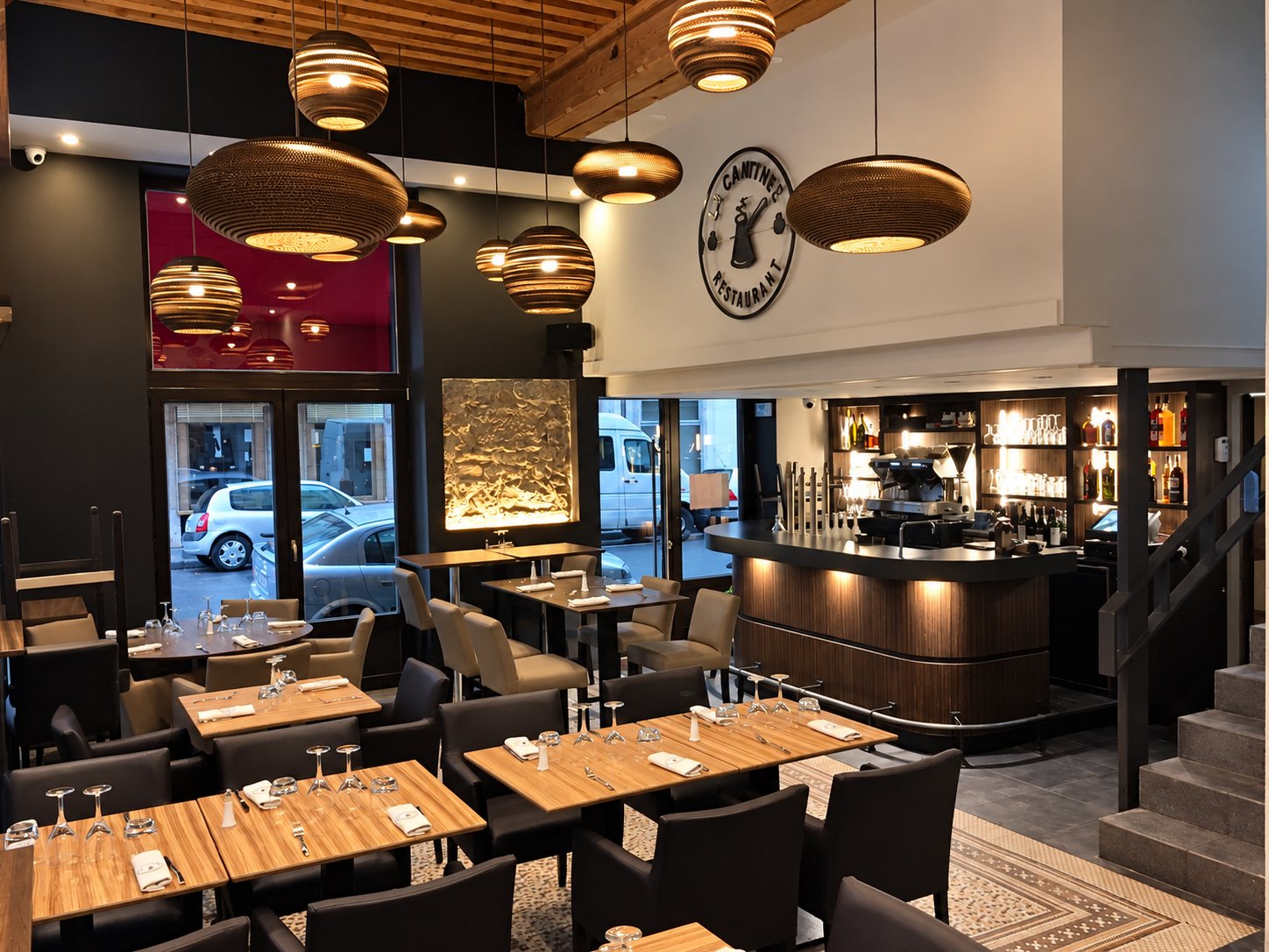Intérieur d'un restaurant aux tables en bois dressées, éclairé par de grandes suspensions en carton, avec un bar et une horloge-enseigne au fond.