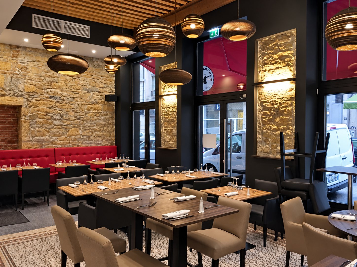 Salle de restaurant aux tables dressées, avec un mur en pierre apparente, une banquette rouge et des suspensions en carton ondulé.