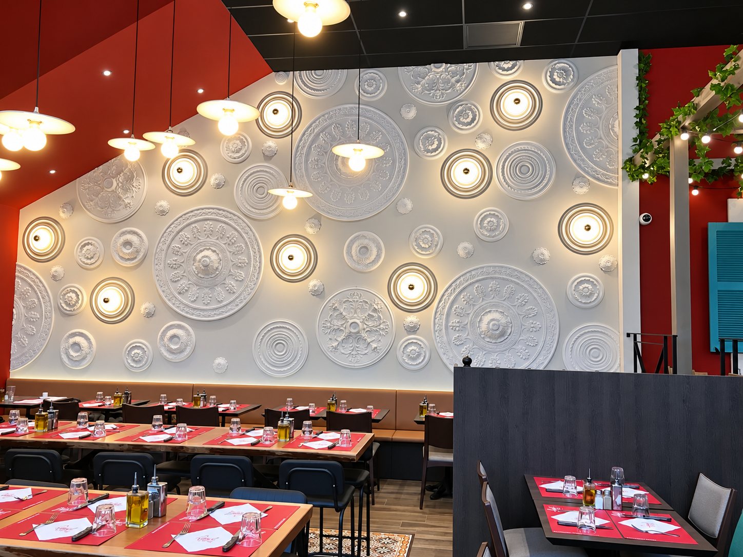 Intérieur d'un restaurant aux tables dressées avec des sets rouges, face à un grand mur blanc décoré de rosaces en plâtre et de lampes rondes.