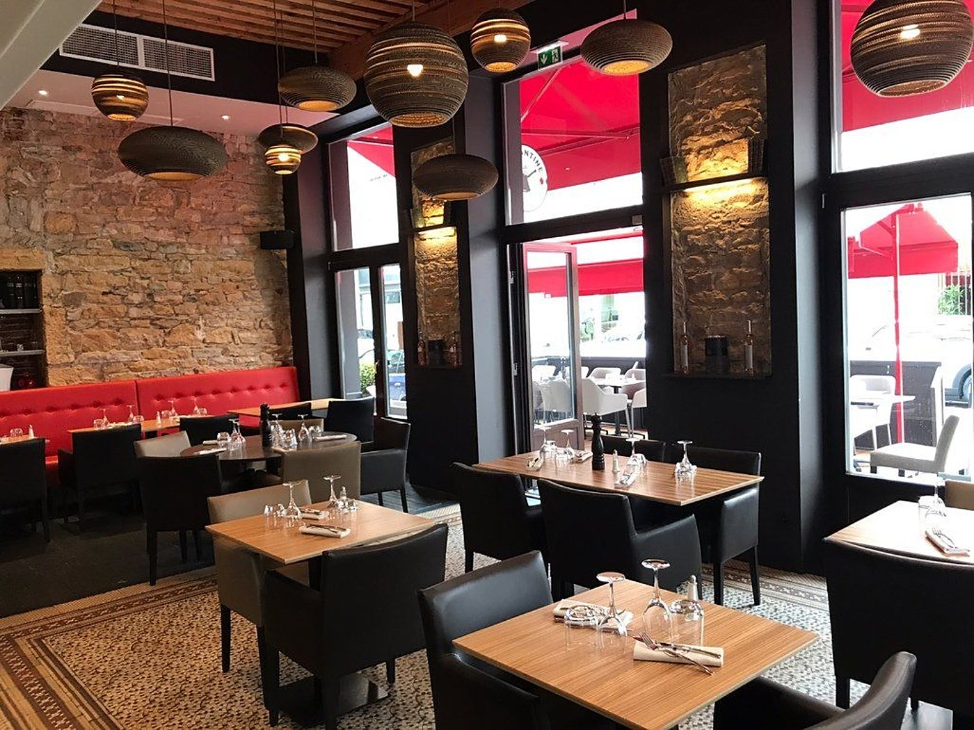 Intérieur d'un restaurant avec tables en bois dressées, fauteuils noirs, banquette rouge, mur en pierre et suspensions en carton ondulé, ouvert sur une terrasse.