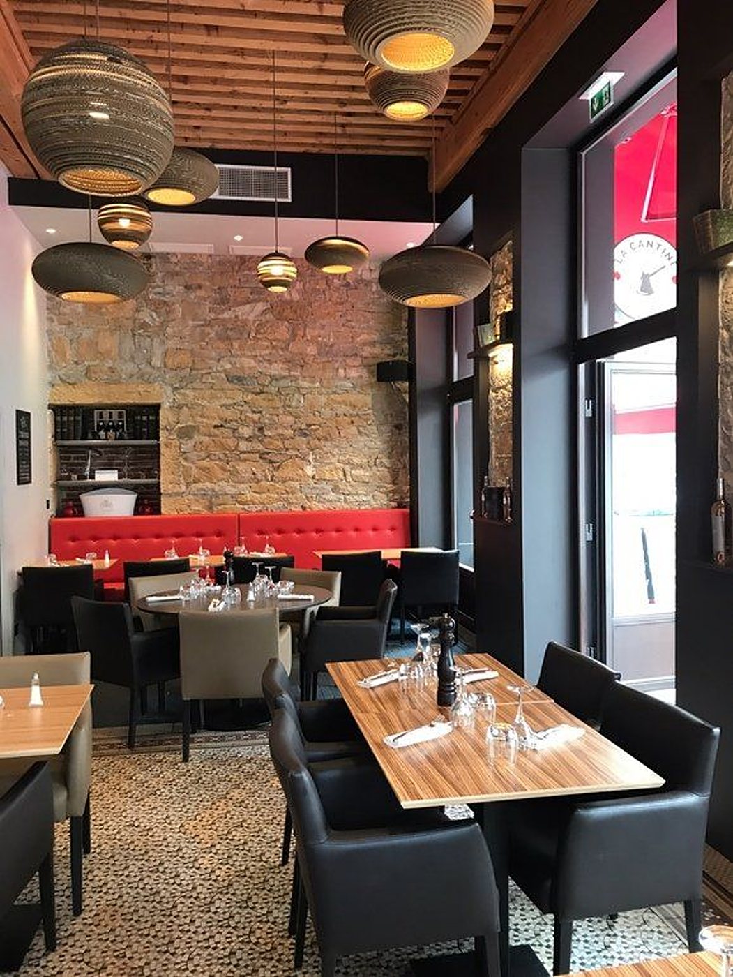 Intérieur d'un restaurant avec un mur en pierre, des banquettes rouges, des tables en bois dressées et des suspensions en carton au plafond.