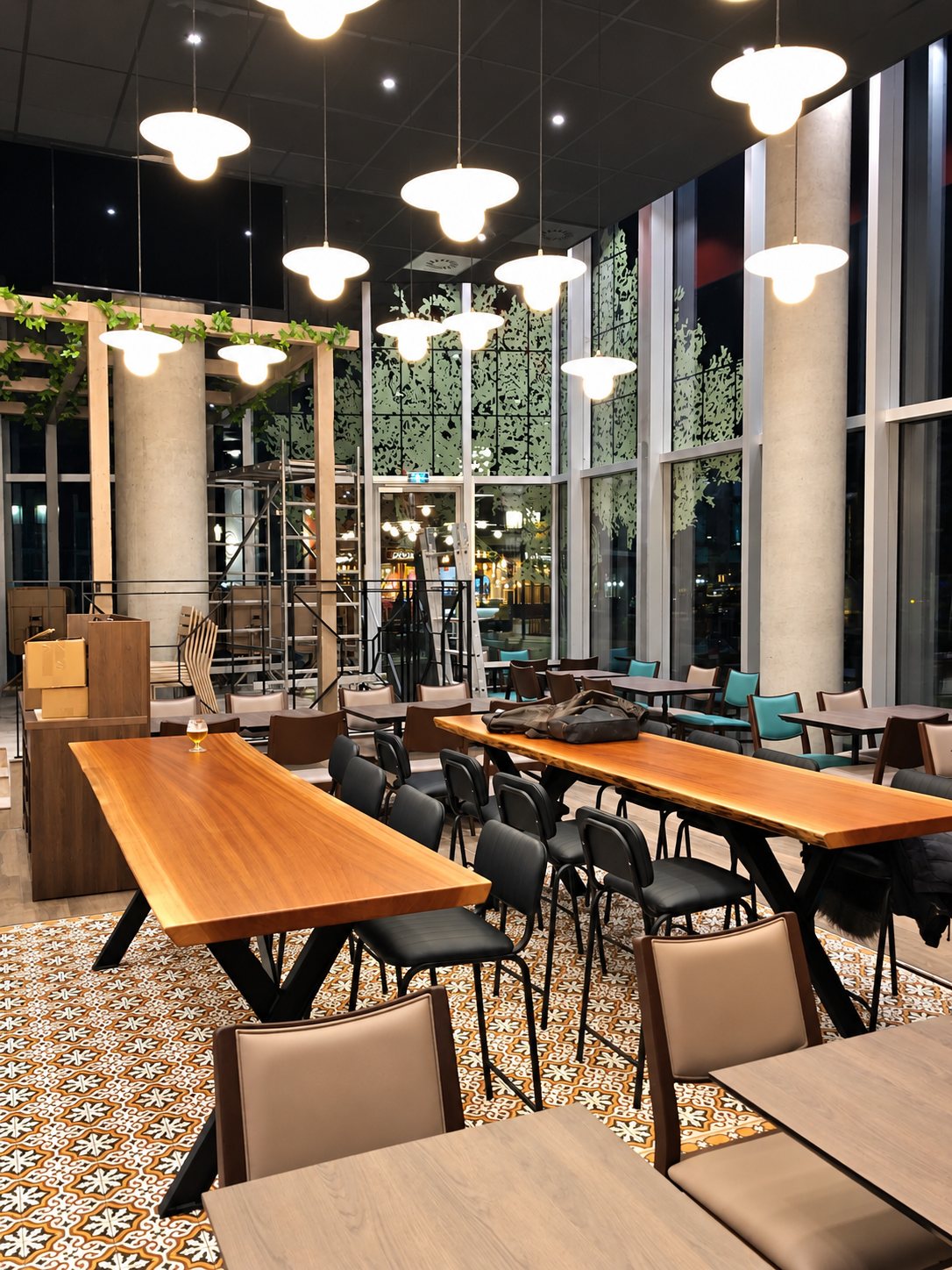 Intérieur d'un restaurant éclairé le soir, avec de longues tables en bois à bord naturel, des chaises noires, un sol en carreaux de ciment et des suspensions blanches.