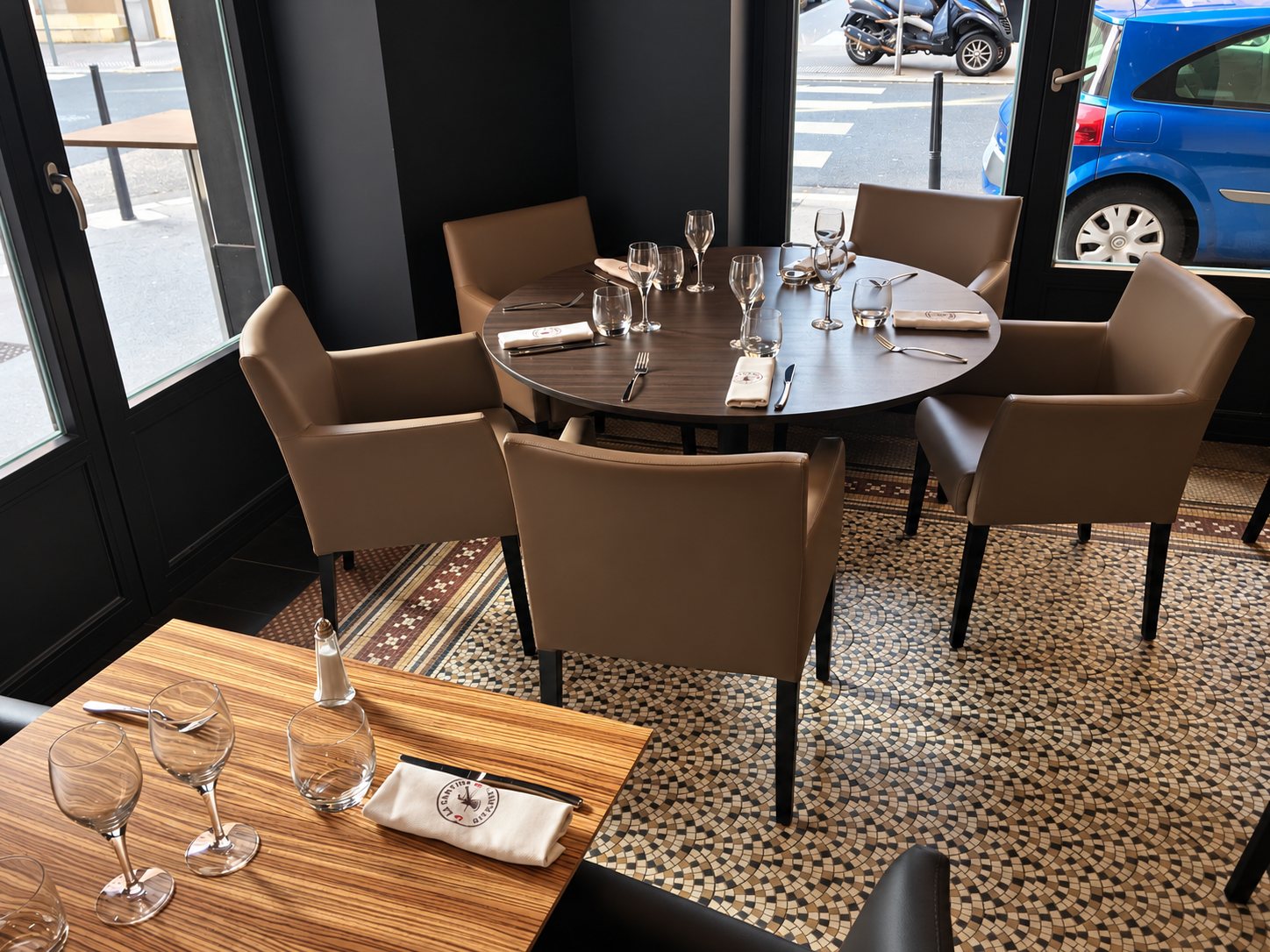 Salle de restaurant lumineuse avec une table ronde et une table en bois dressées, entourées de fauteuils beiges sur un sol en mosaïque.