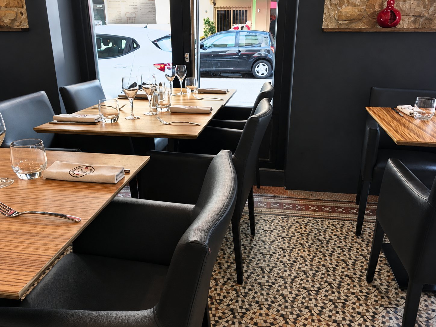 Intérieur d'un restaurant avec des tables en bois dressées, des fauteuils en cuir sombre et un sol en mosaïque devant une grande vitrine donnant sur la rue.